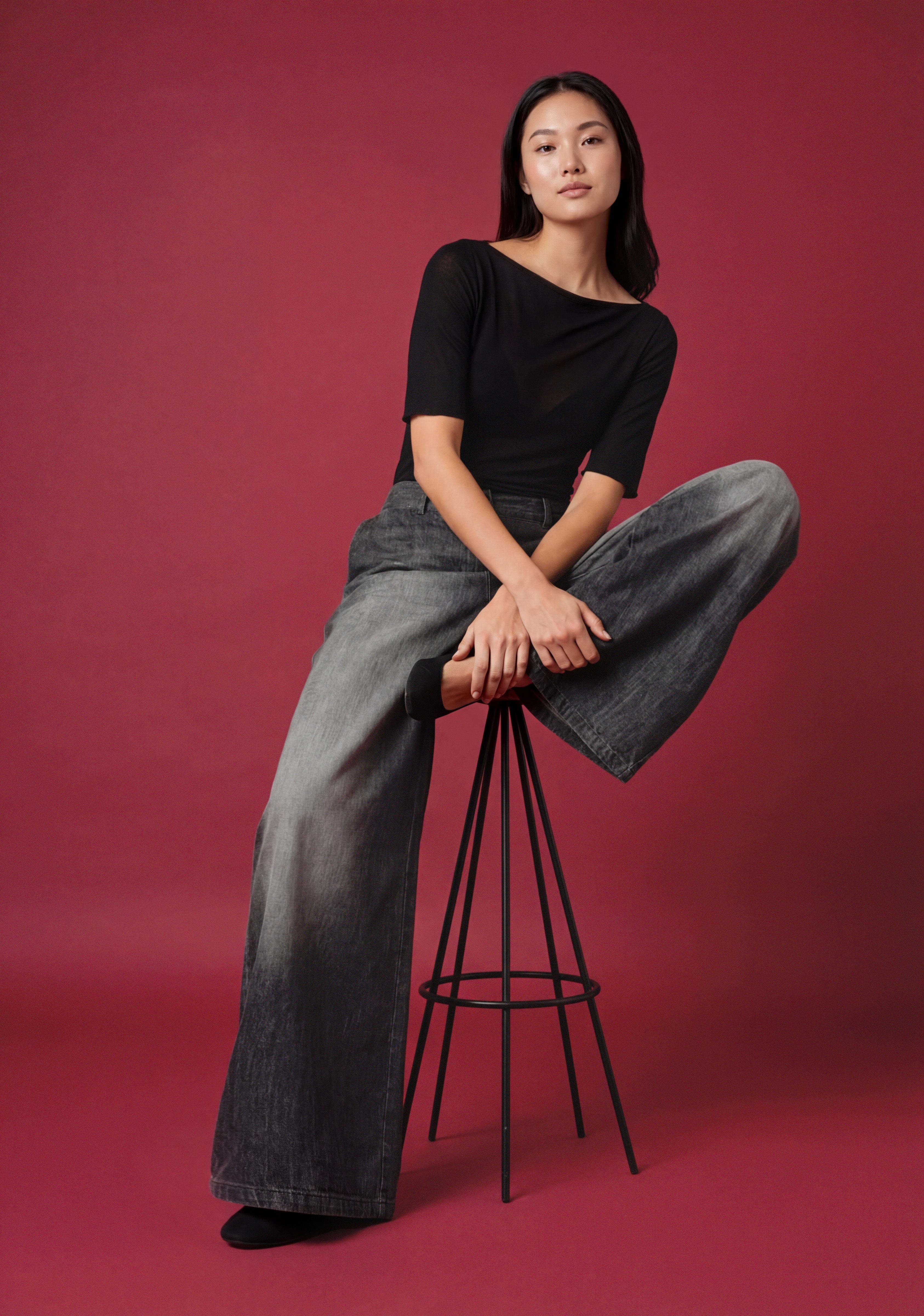 Model sitzt auf einem schwarzen Hocker vor rotem Hintergrund und trägt ein schwarzes Oberteil mit weiter grauer Jeans.