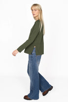 Woman wearing olive green HAEIN Clean Jersey long sleeve with U-boat neckline and side slits paired with blue wide-leg jeans and brown boots.