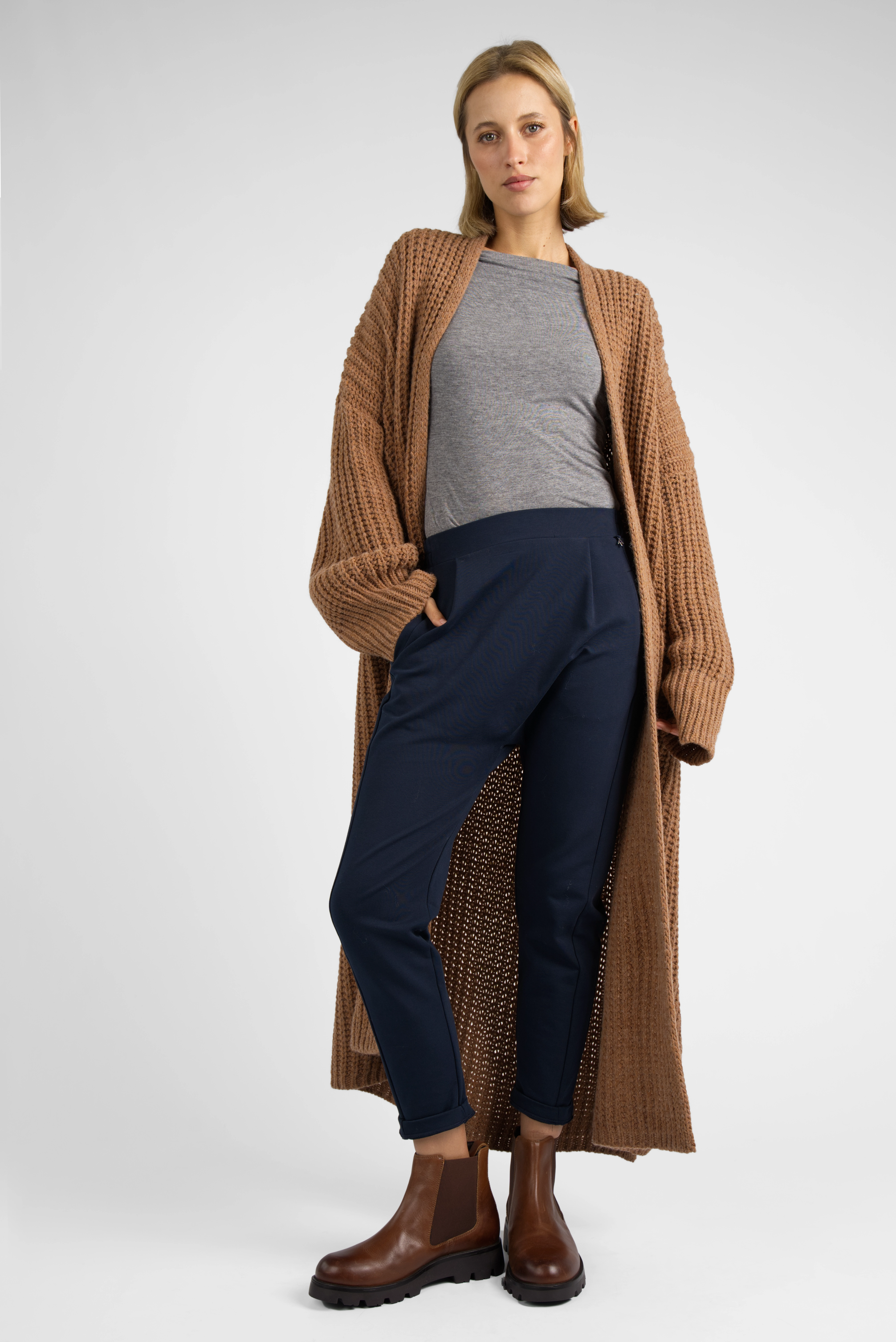 Frau trägt HANNA.K Tapered Sweathose in Navy mit elastischem Bund und schmal zulaufendem Bein, kombiniert mit grauem Shirt, braunem Strickmantel und braunen Stiefeln.