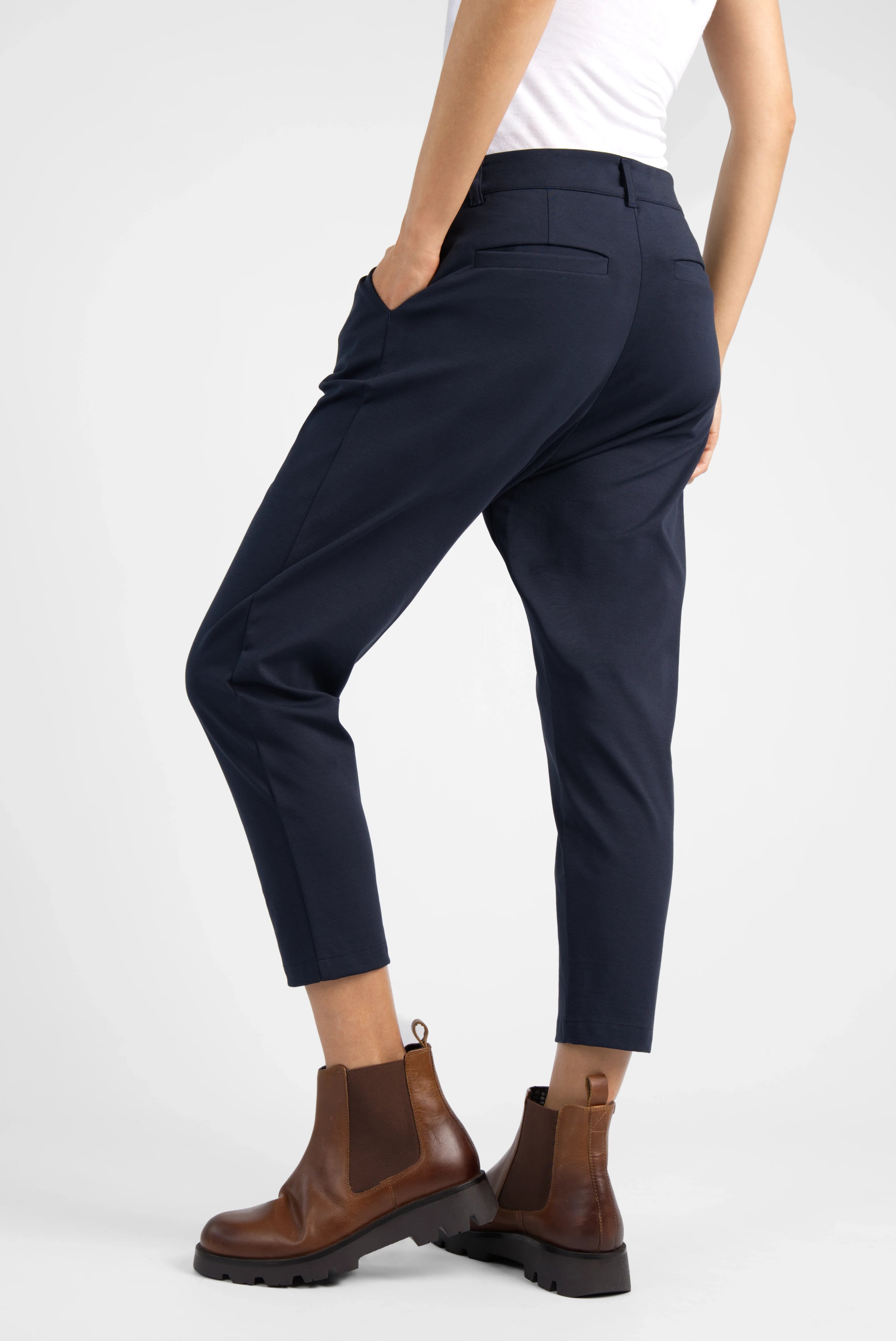 Damen cropped HEELENA OG Tapered Pants in navy, schmal zulaufend, mit Bundfalten und Reißverschluss, kombiniert mit braunen Stiefeln.