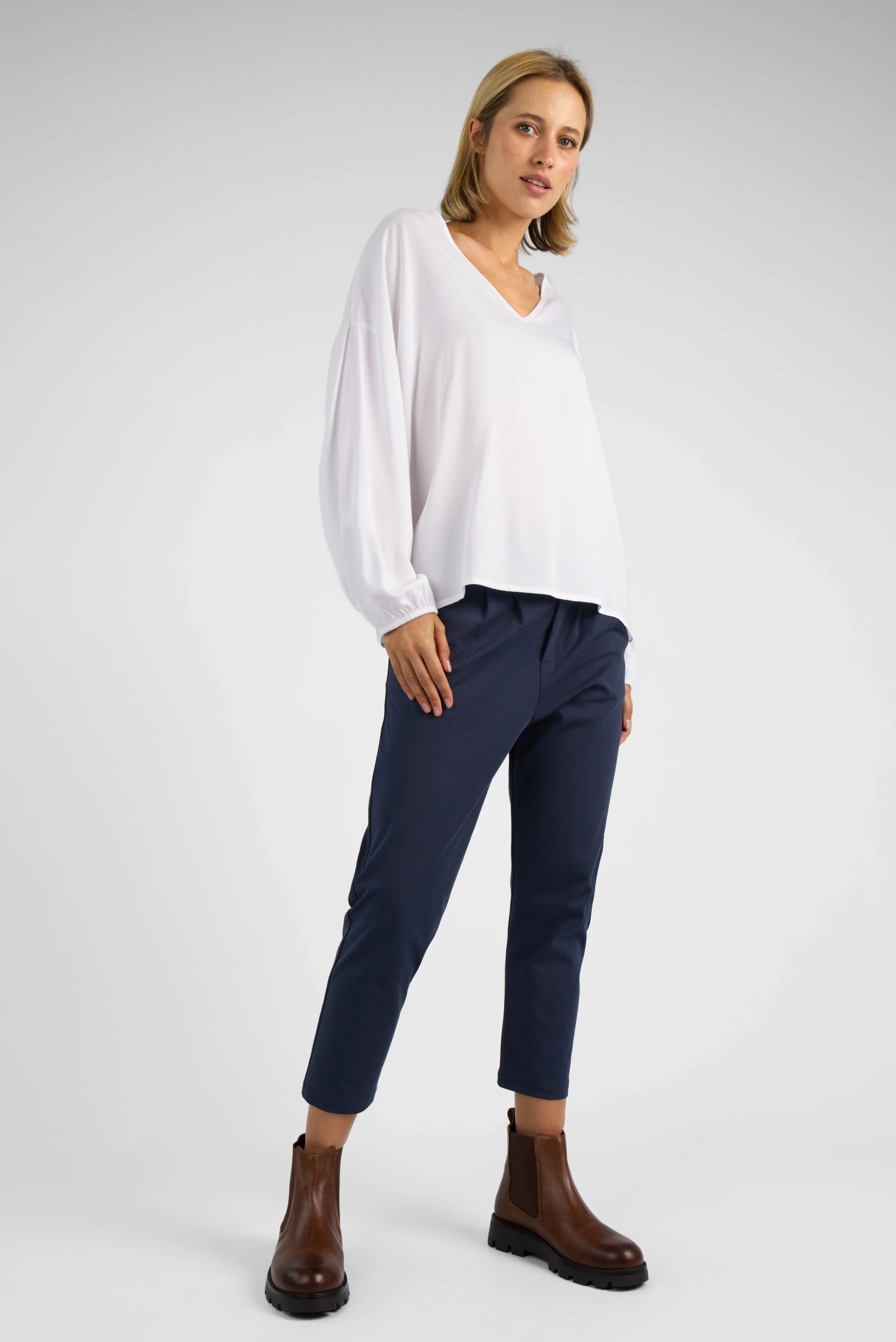 Frau trägt dunkelblaue HEELENA OG Tapered Pants cropped mit weißem V-Ausschnitt Pullover und braunen Stiefeln vor grauem Hintergrund