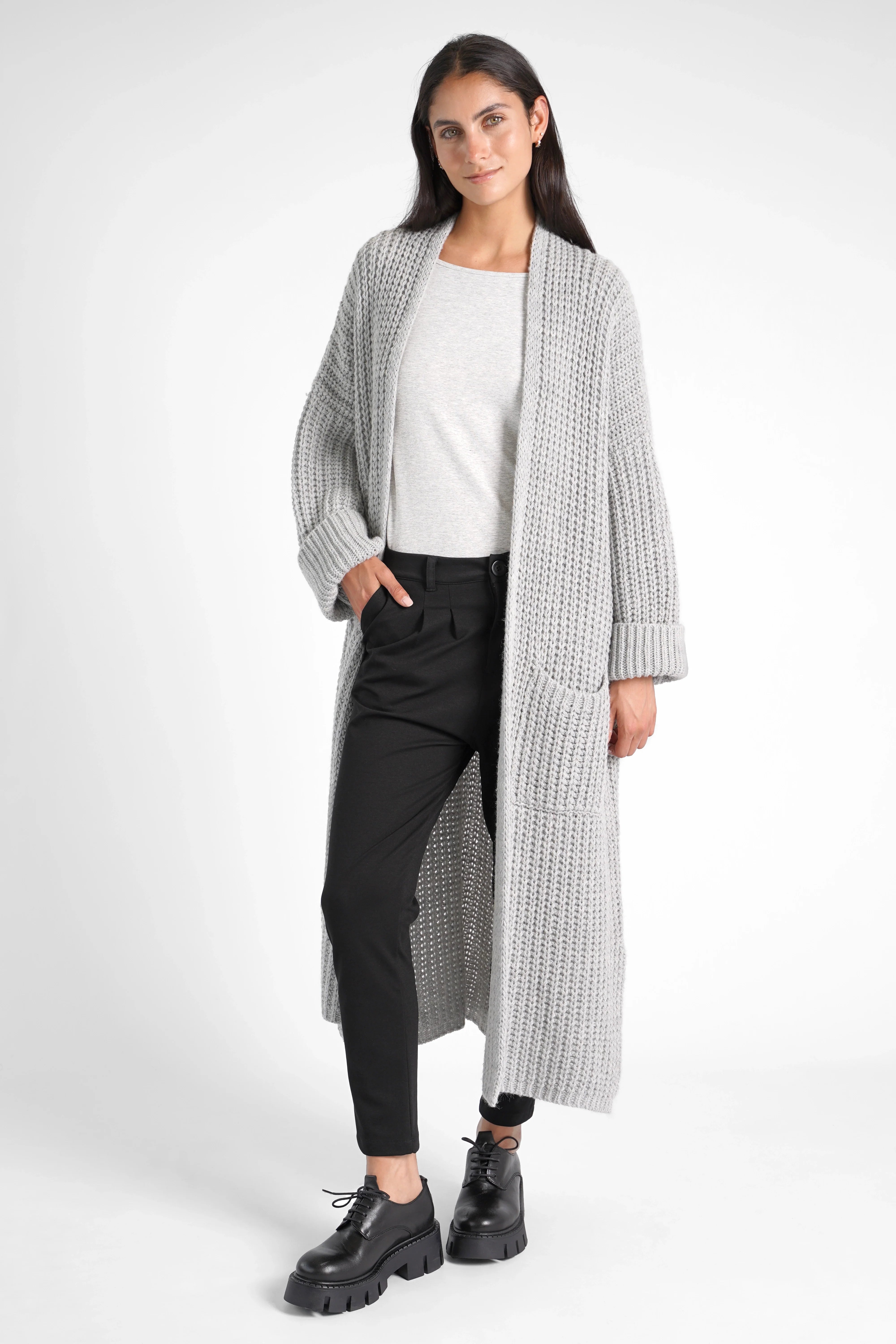 Frau trägt schwarze HEELENA OG Tapered Pants mit schmal zulaufendem Bein und normaler Bundhöhe kombiniert mit grauem Longcardigan und schwarzen Schnürschuhen vor weißem Hintergrund.