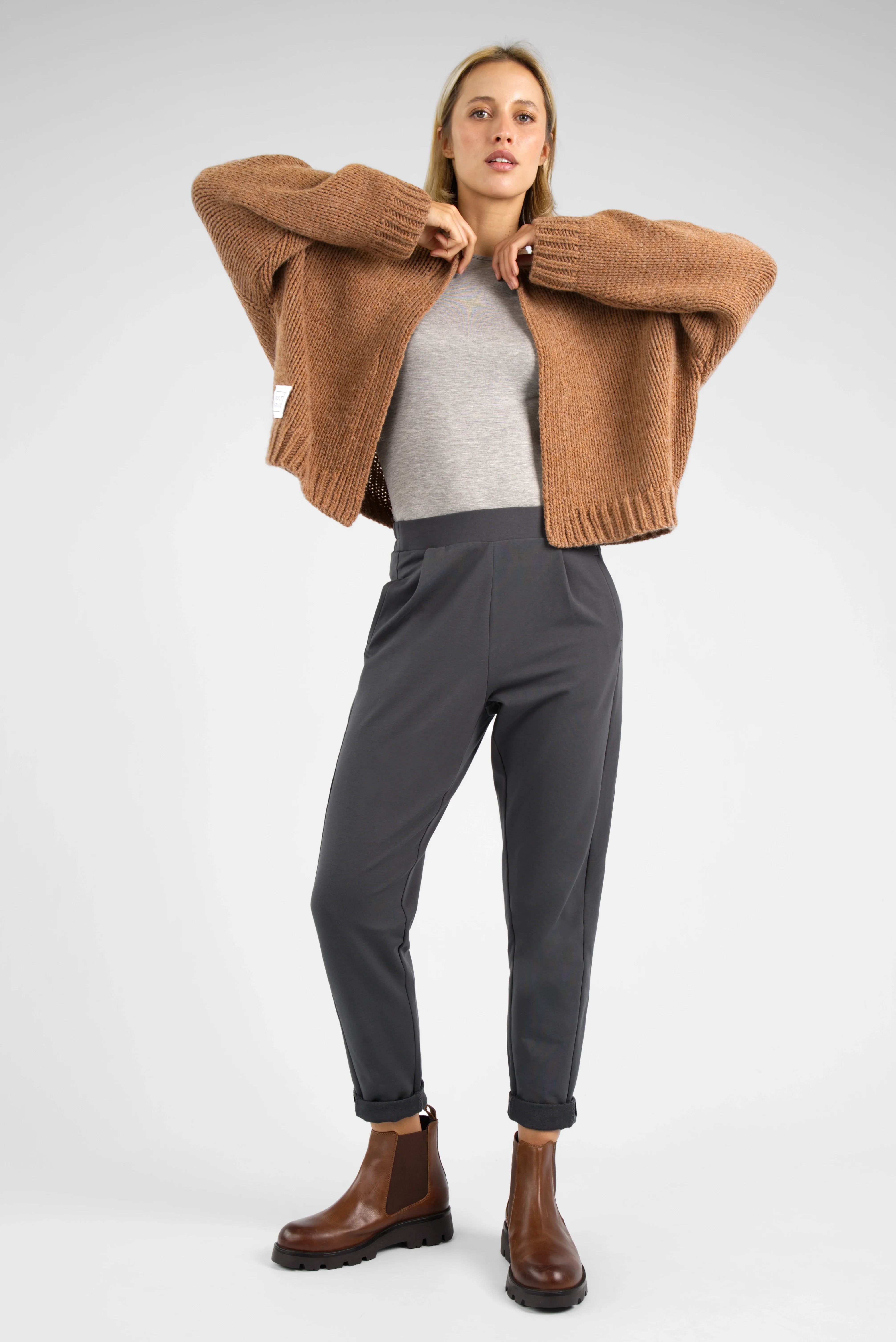 Damen trägt HILLA.K Jerseyhose in dunklem Grau mit mittelhohem Bund und leicht zulaufendem Bein kombiniert mit brauner Strickjacke und braunen Stiefeln