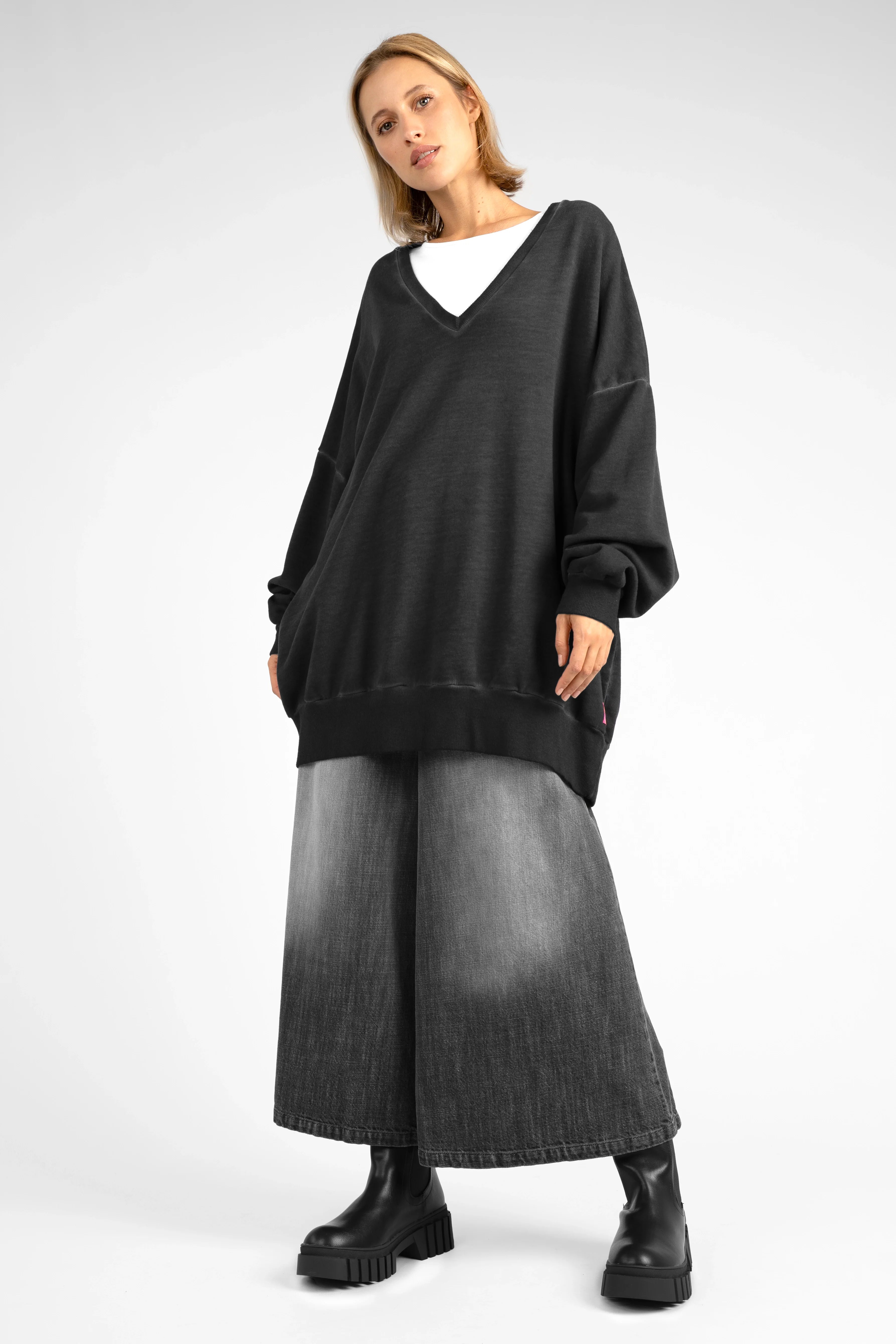 Woman wearing black oversized IMIKO Studio long-sweater with V-neck and dropped shoulders, paired with wide leg jeans and black boots.