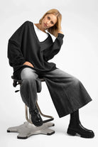 Woman wearing oversized black IMIKO Studio Long-Sweater with deep V-neck and dropped shoulders, paired with wide-leg jeans and black boots, seated on a modern swivel chair against a white background.