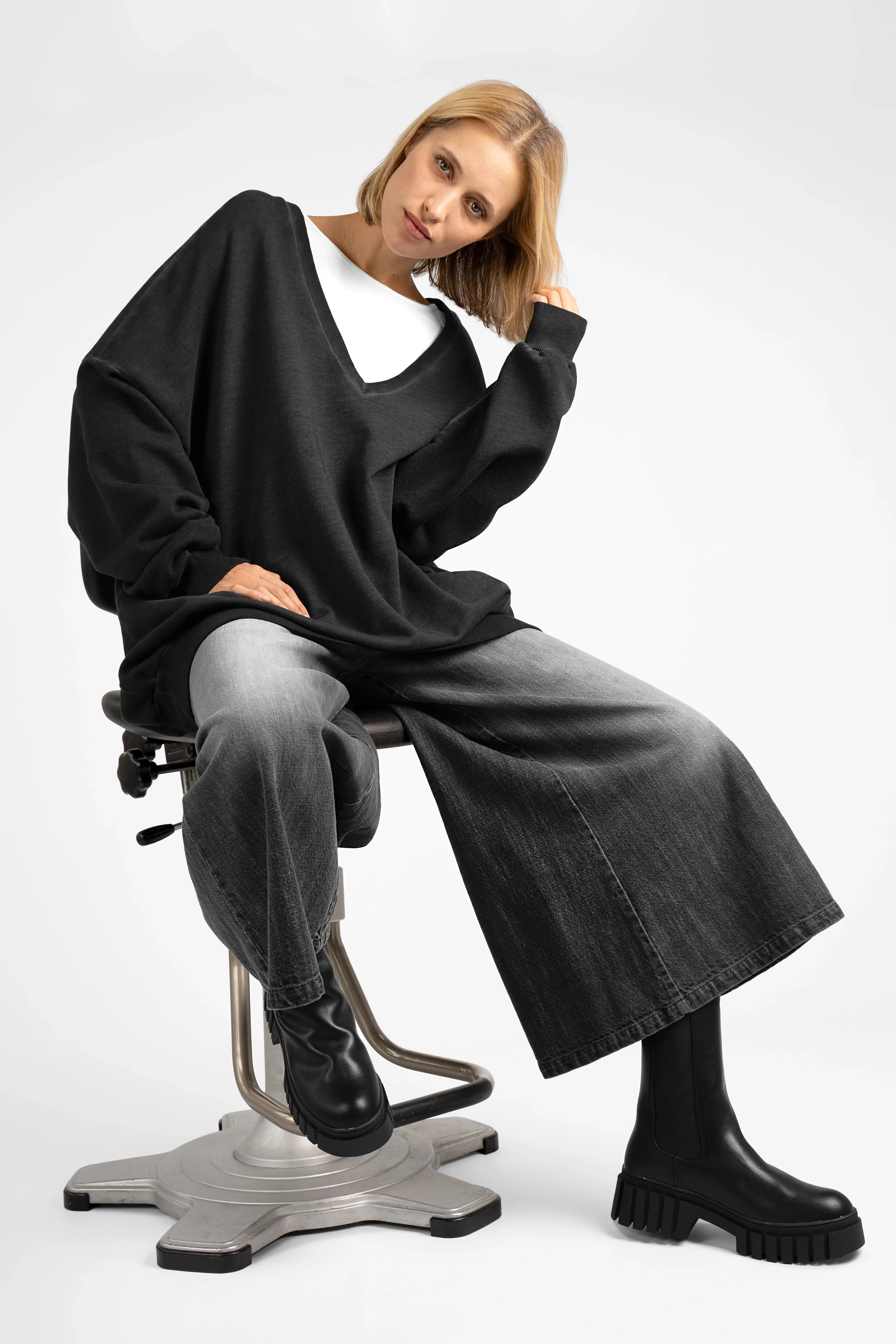 Woman wearing oversized black IMIKO Studio Long-Sweater with deep V-neck and dropped shoulders, paired with wide-leg jeans and black boots, seated on a modern swivel chair against a white background.