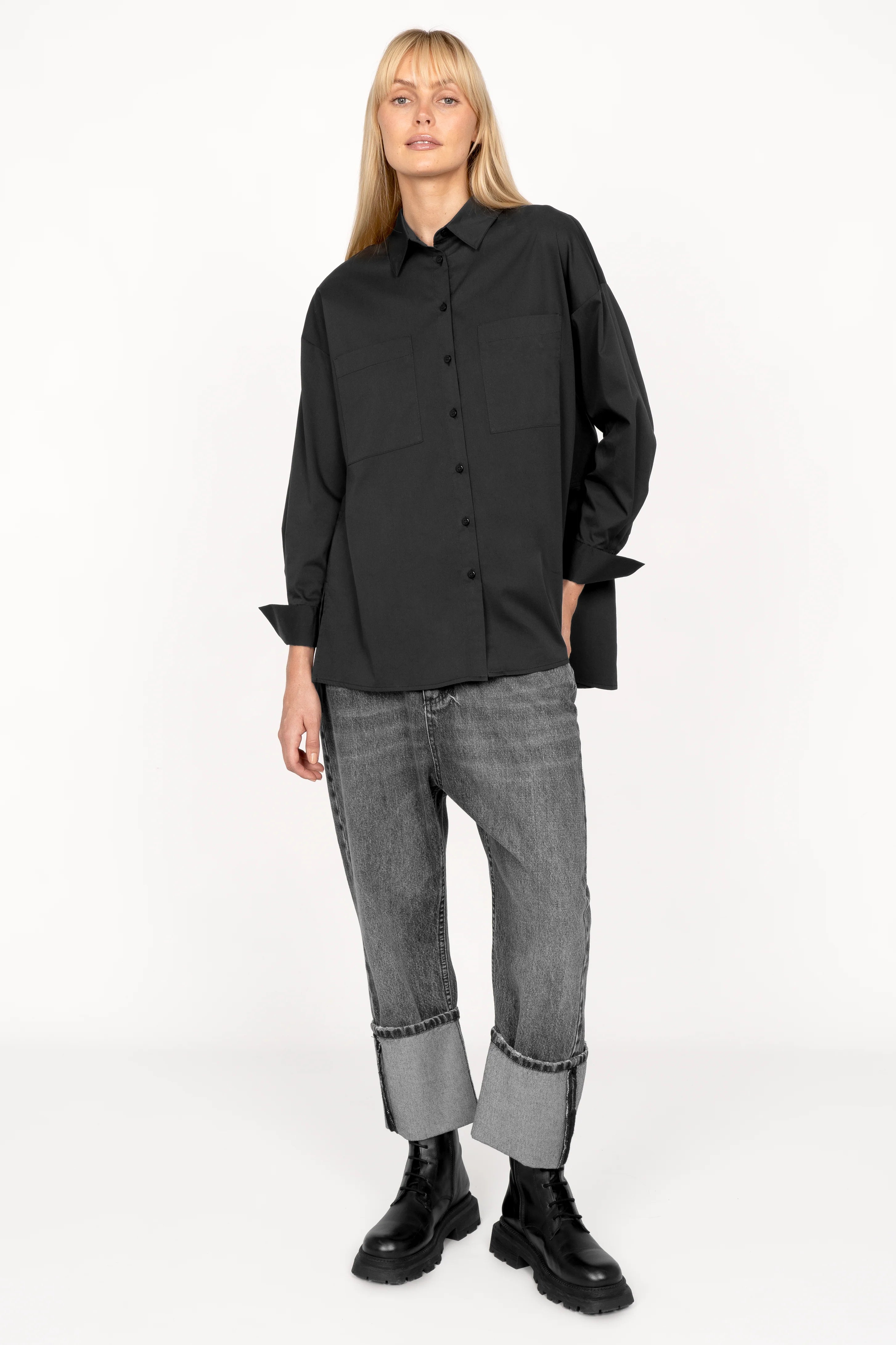 Woman wearing black LEA.K loose fit blouse with hem collar, chest pockets, wide buttoned cuffs, and longer trapezoidal back paired with cuffed jeans and black boots