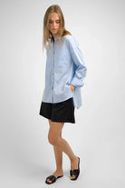 Woman wearing light blue LEA.K Loose Fit blouse with long sleeves and black shorts standing against a plain gray background