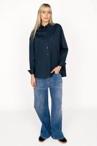 Woman wearing navy blue LEA.K loose fit blouse with button-up front, chest pockets, wide cuffs, and longer back paired with blue jeans
