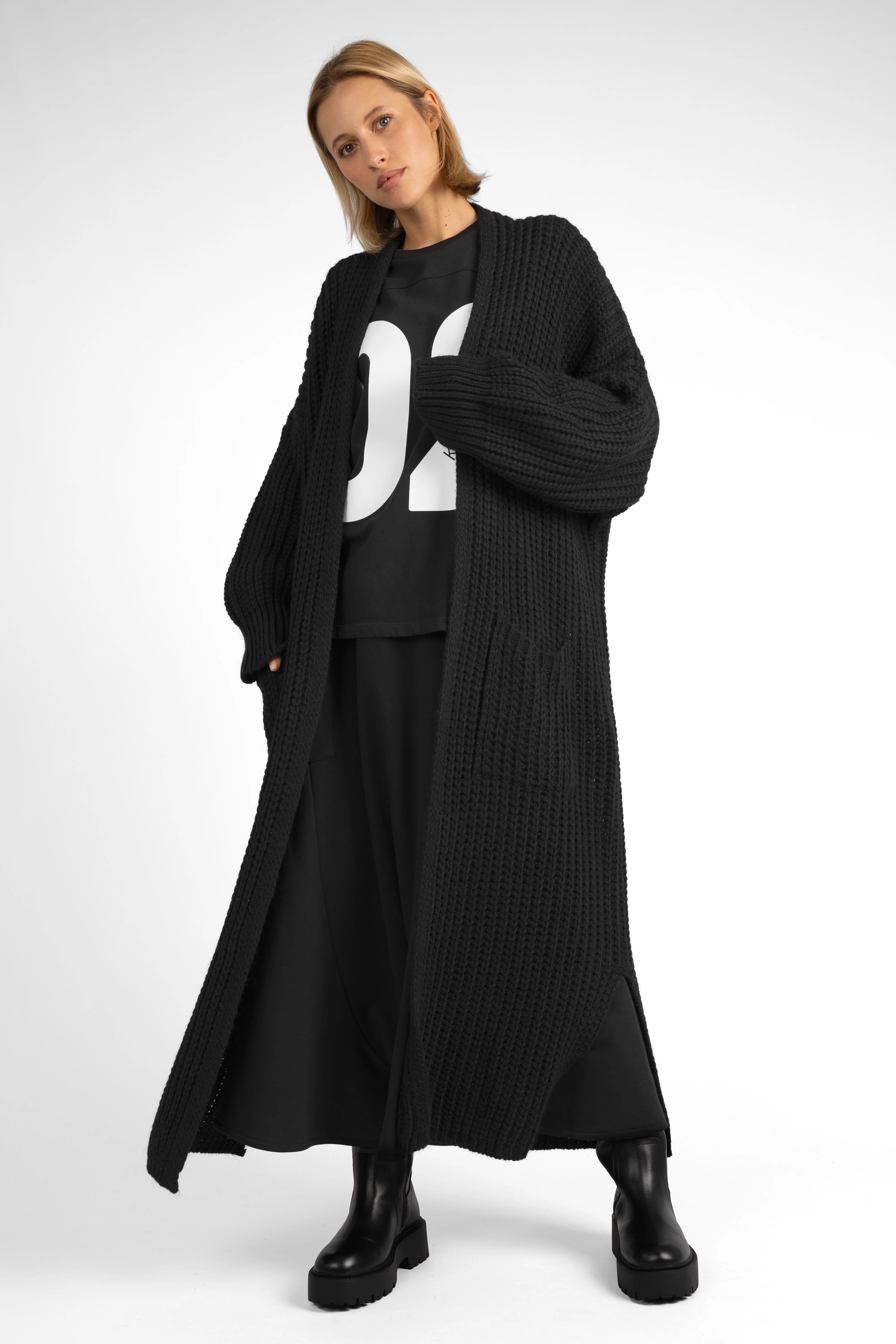 Woman wearing a black oversized LOLA Strickcardigan with relaxed fit and open front, styled with black pants and boots.