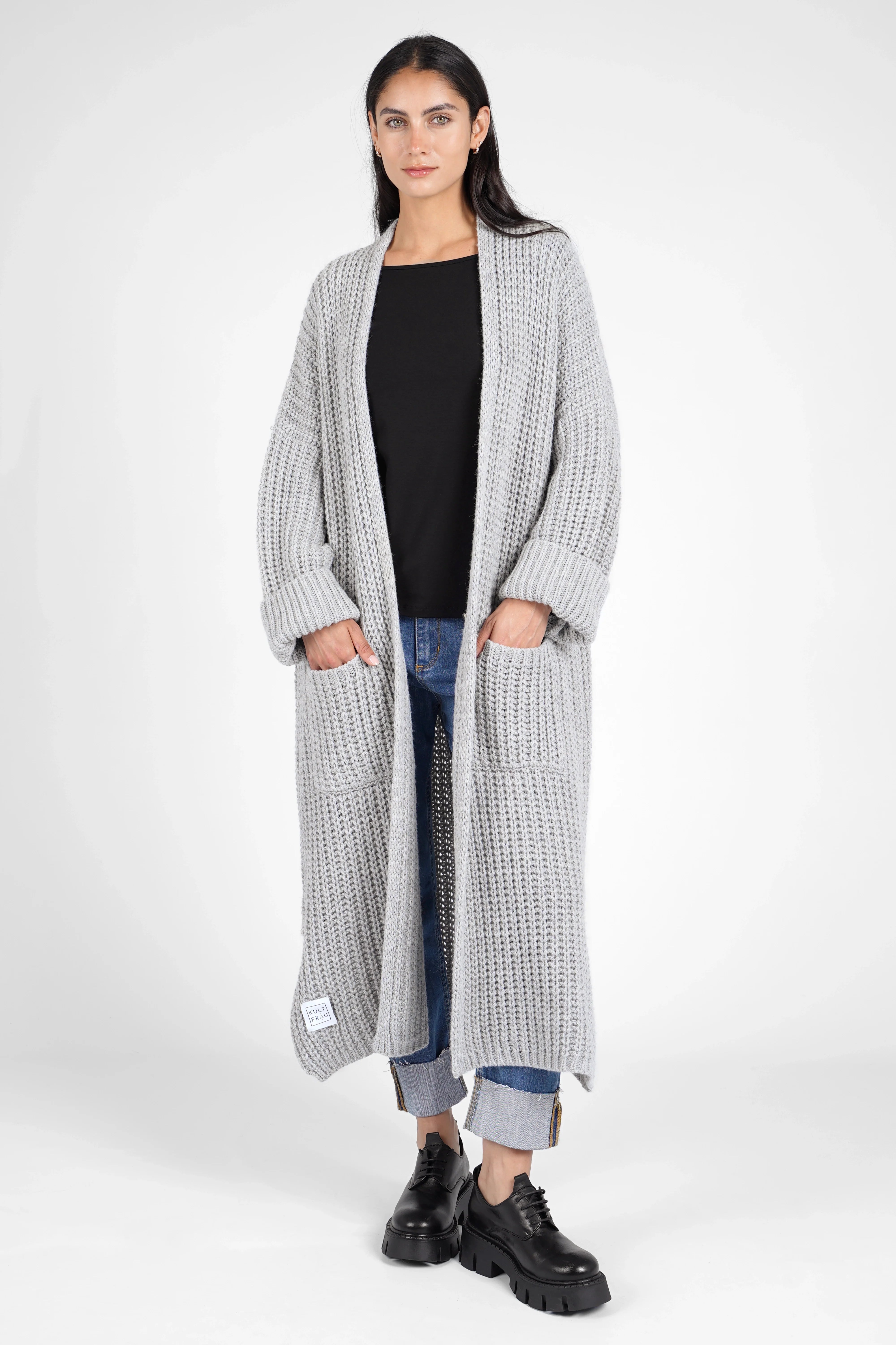Woman wearing a grey LOLA Strickcardigan oversized knit cardigan with rolled-up sleeves and front pockets over a black top and blue jeans