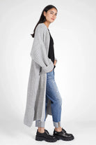 Woman wearing gray oversized LOLA Strickcardigan with rolled sleeves, paired with black top, blue jeans, and black chunky shoes.