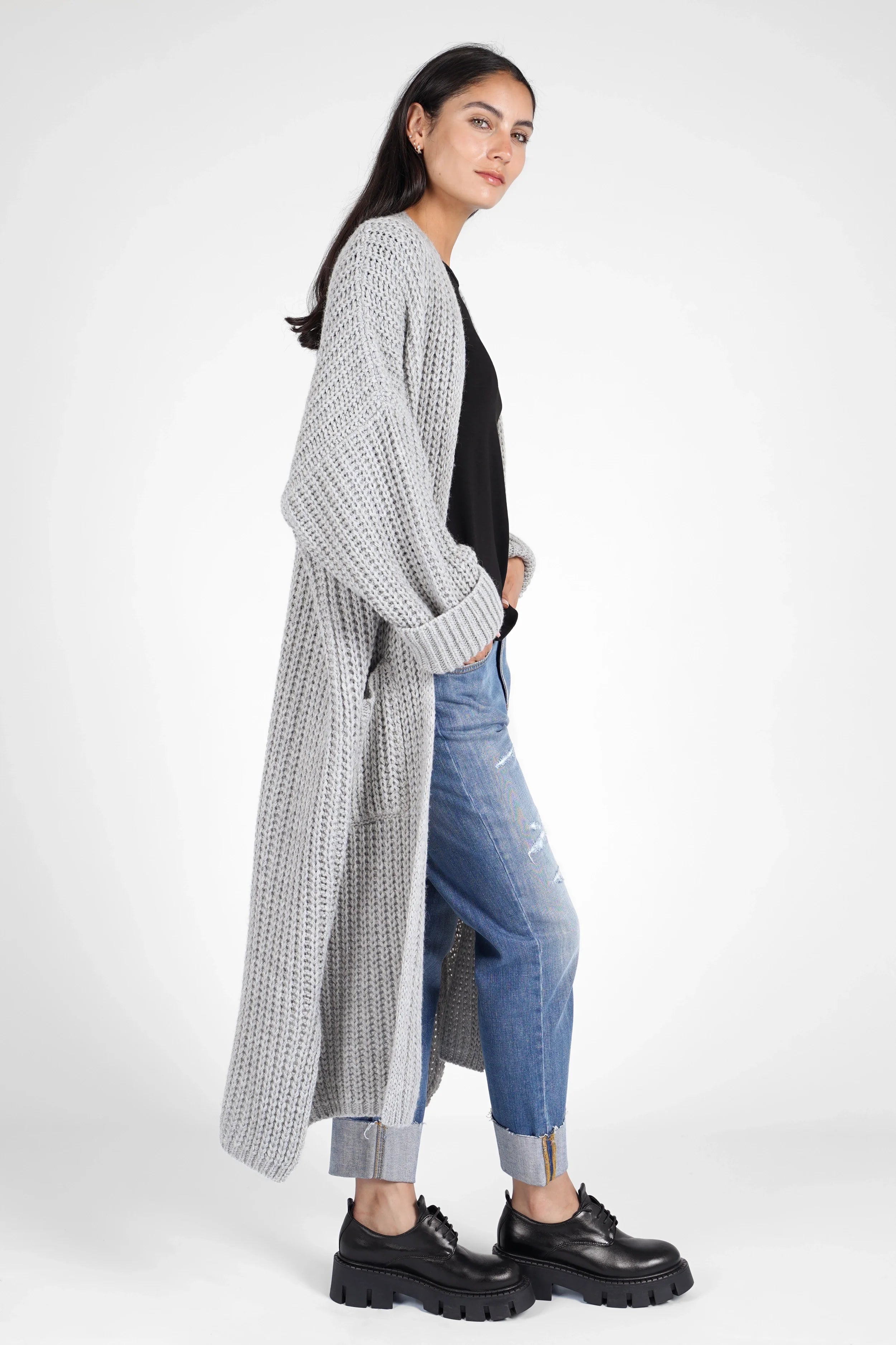 Woman wearing gray oversized LOLA Strickcardigan with rolled sleeves, paired with black top, blue jeans, and black chunky shoes.