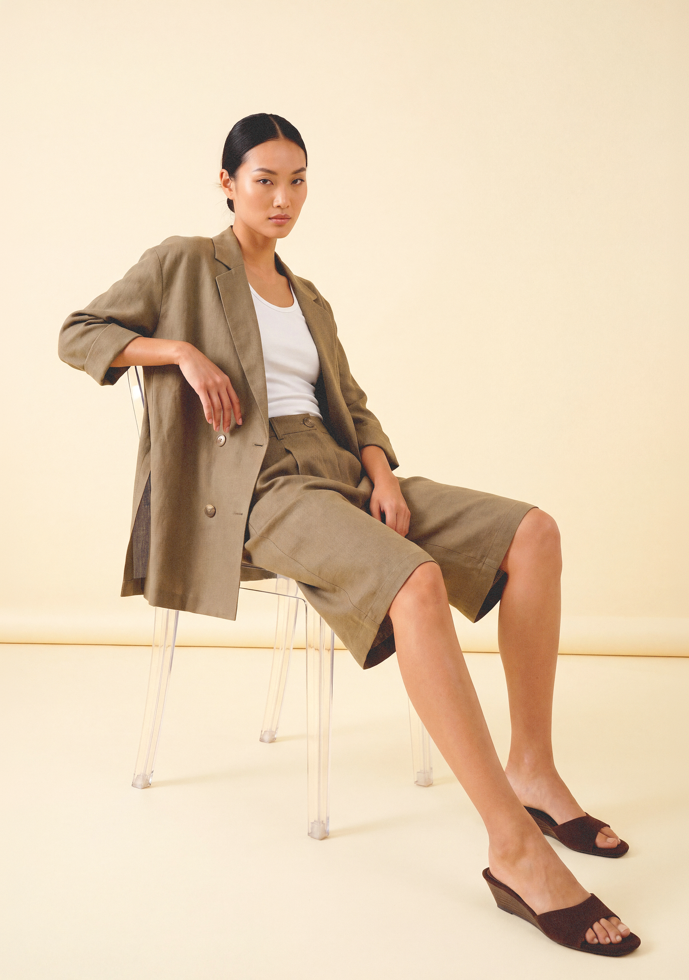 Model im beigefarbenen Leinen-Zweiteiler mit Blazer und Bermuda-Shorts, minimalistisch inszeniert vor neutralem Hintergrund.