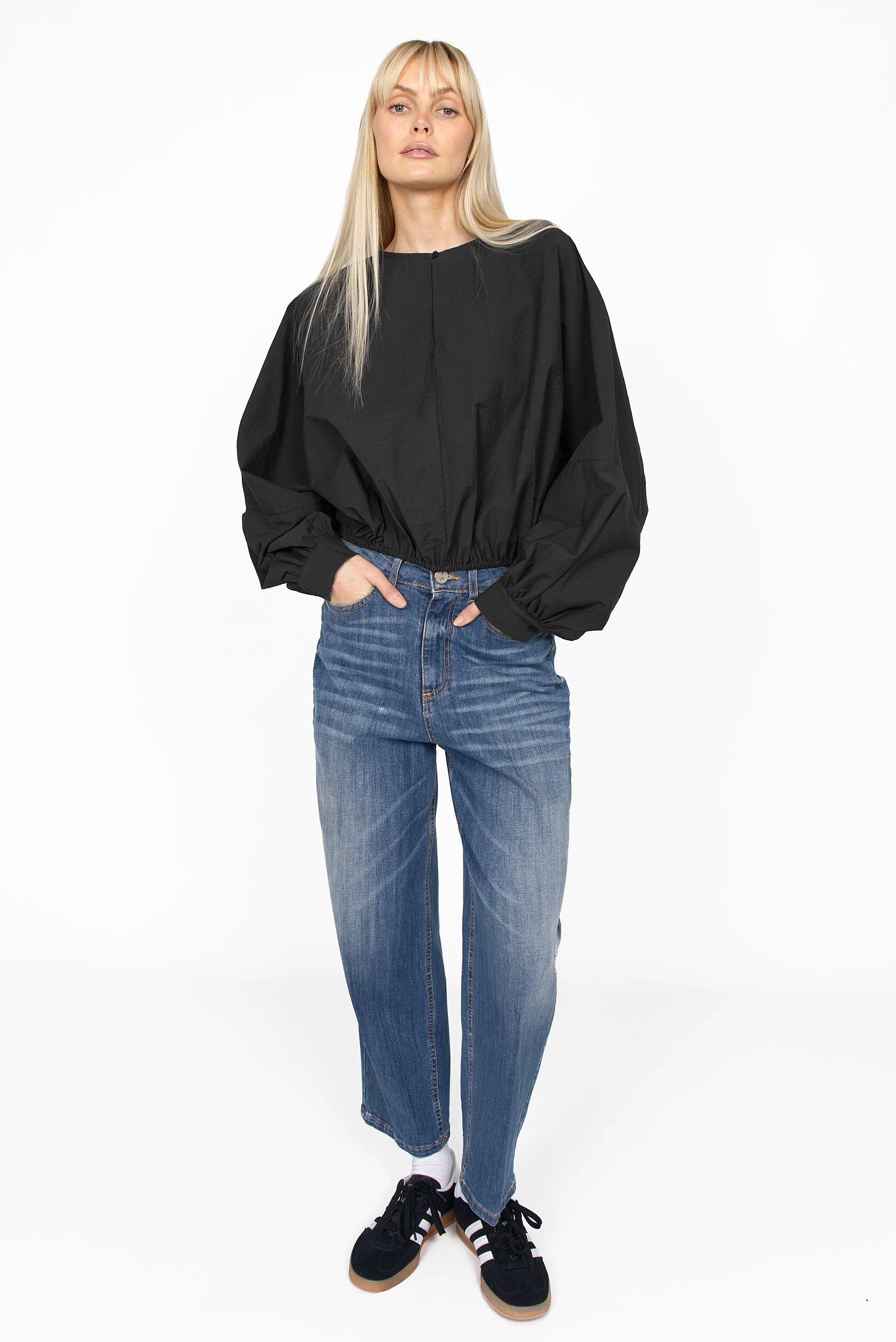 Woman wearing black MILLIE Crinkle Bluse with balloon sleeves and elastic hem paired with blue jeans and black sneakers.