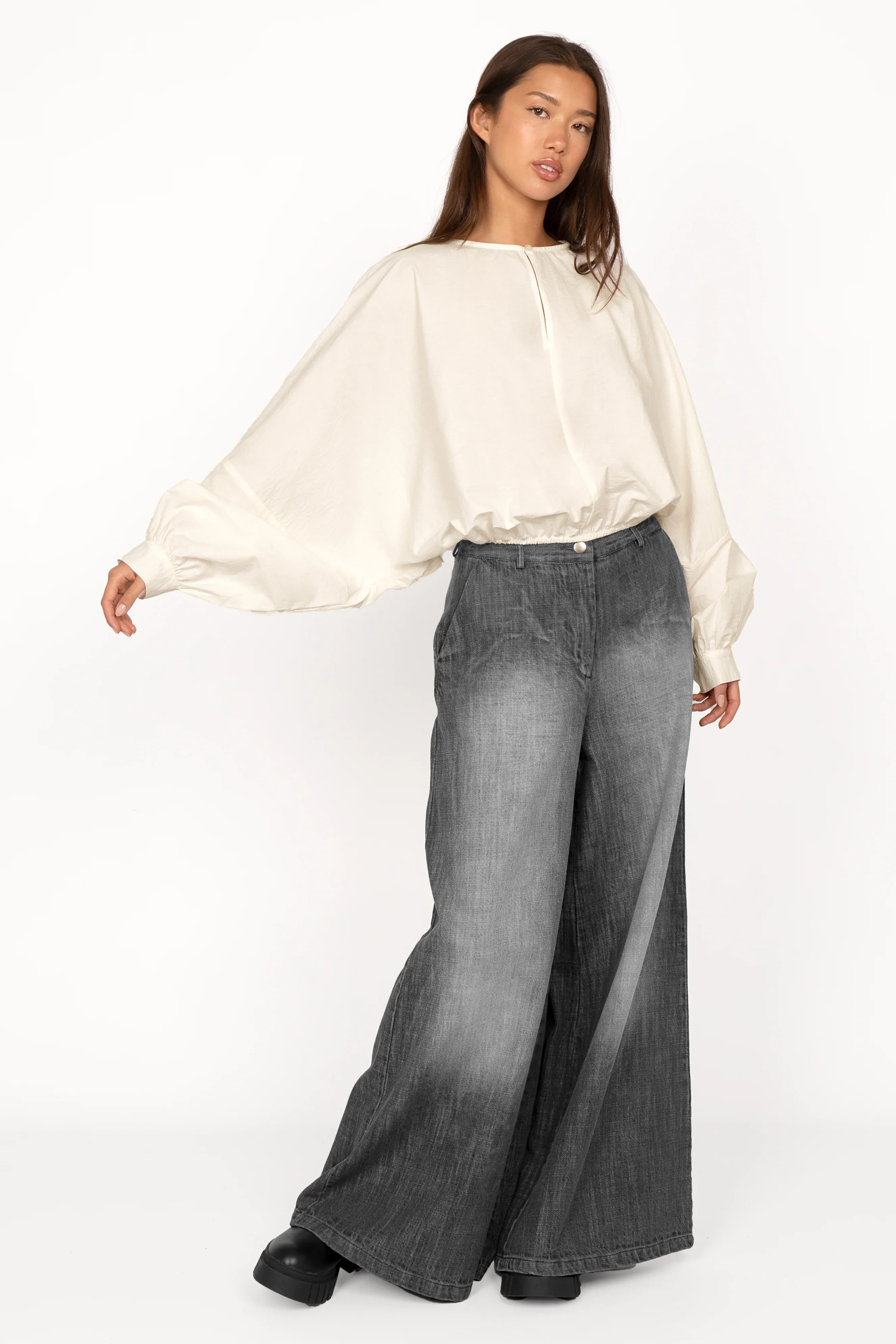 Model wearing a loose fit MILLIE Crinkle Bluse with sculptural balloon sleeves, pearl button neck detail, elastic waistband, and cropped length, paired with wide-leg gray pants.