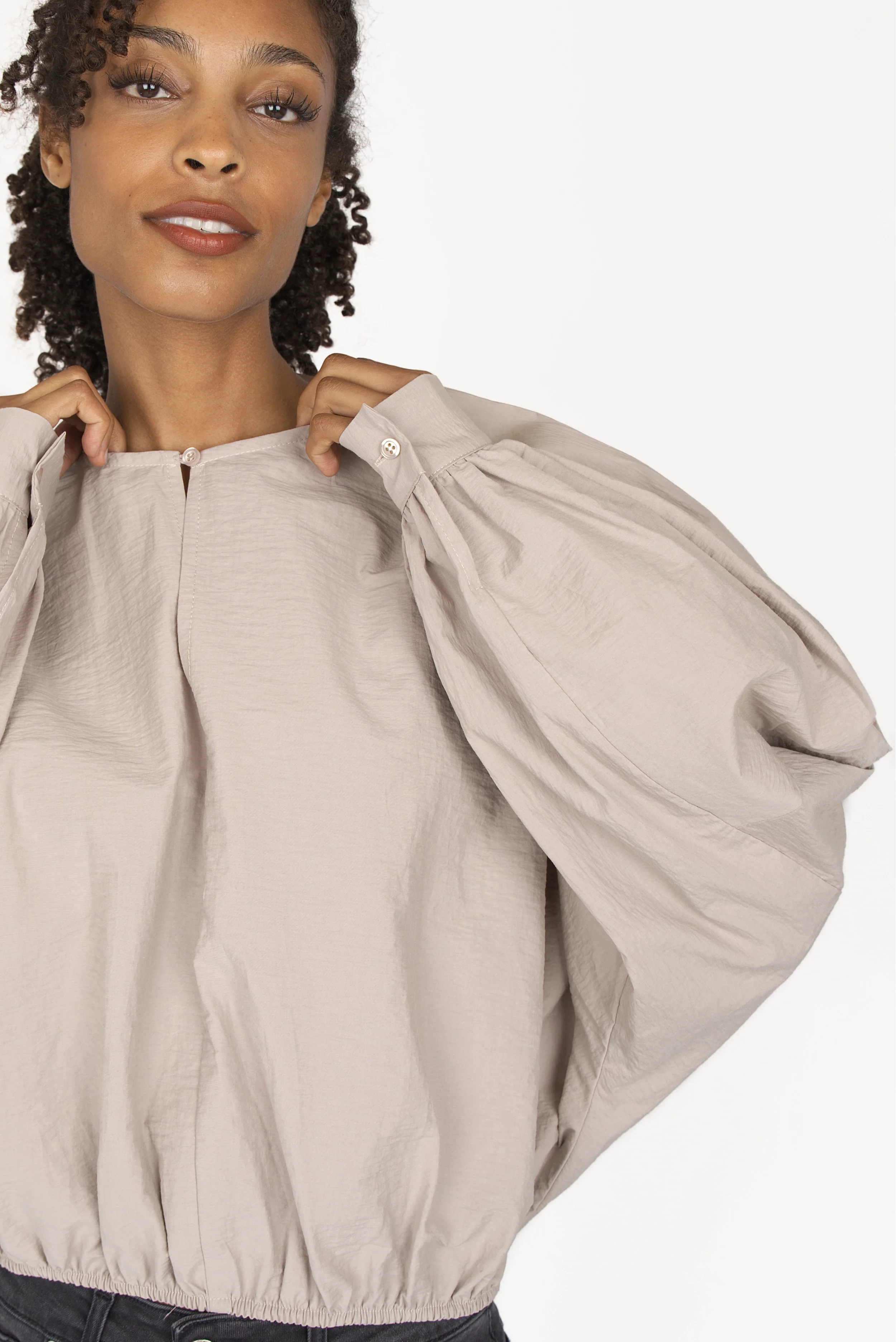 Woman wearing a loose-fit MILLIE Crinkle Bluse with sculptural balloon sleeves, elastic waistband, and pearl button at neckline in beige cotton blend.