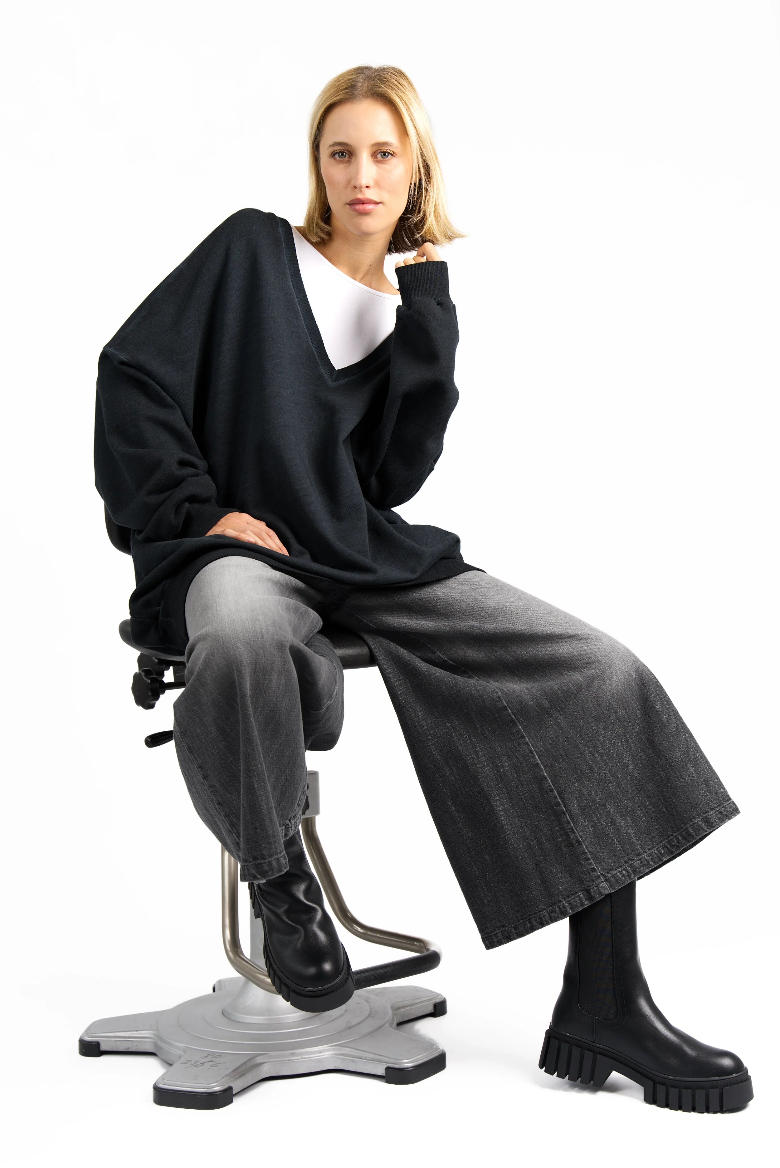 Woman wearing NEDITA Cropped Wide Leg Hose in washed black with gradient, paired with oversized black sweater and black boots sitting on stool against white background