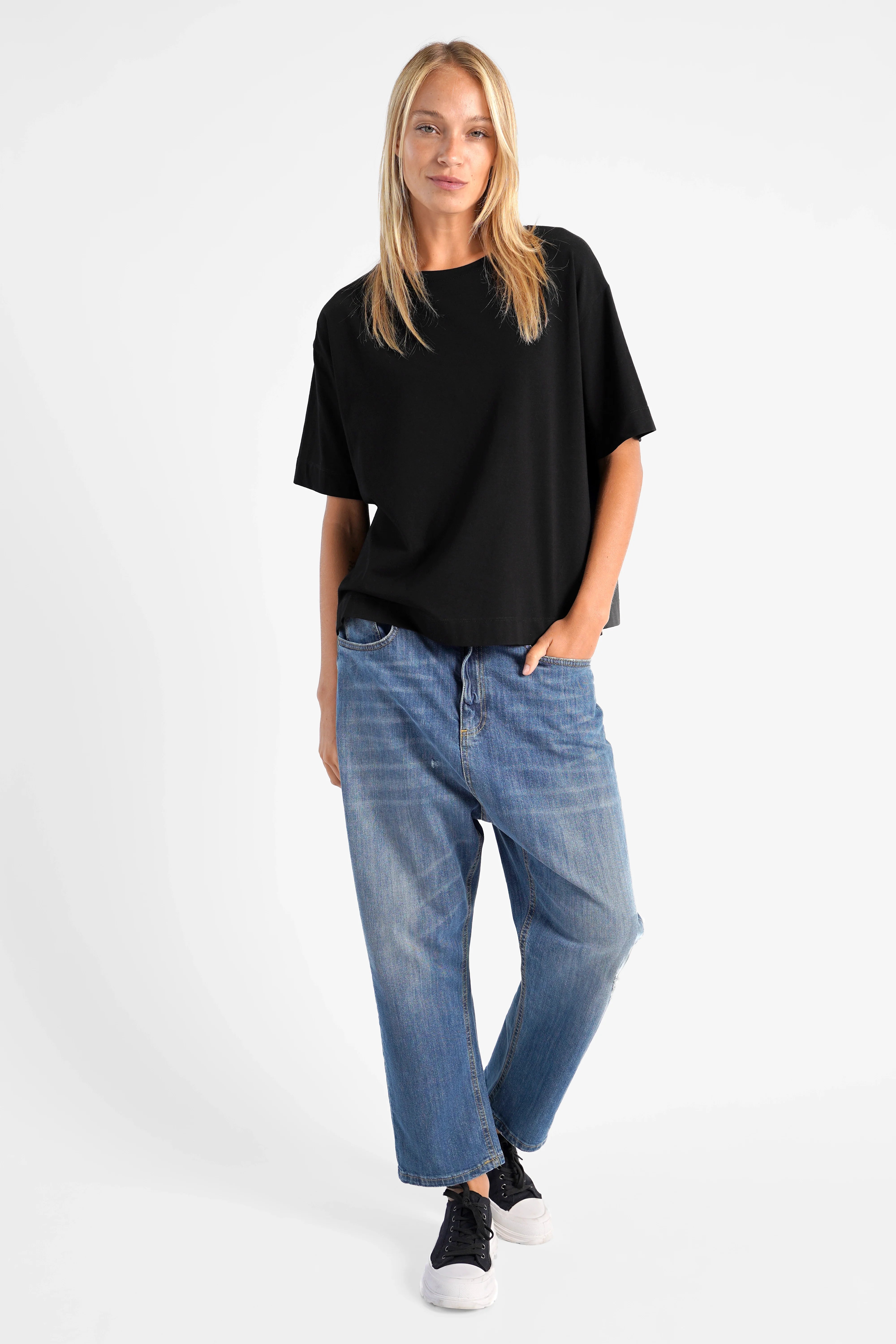 Frau trägt schwarzes NITI.K Shirt aus elastischem Baumwolljersey mit kastiger Passform, überschnittenen Schultern und seitlichen Schlitzen, kombiniert mit blauen Jeans und schwarzen Sneakern.