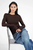 Woman wearing brown NJELI Longsleeve with round neckline, extra long sleeves with thumbholes, and slim fit paired with blue jeans sitting on metal chair