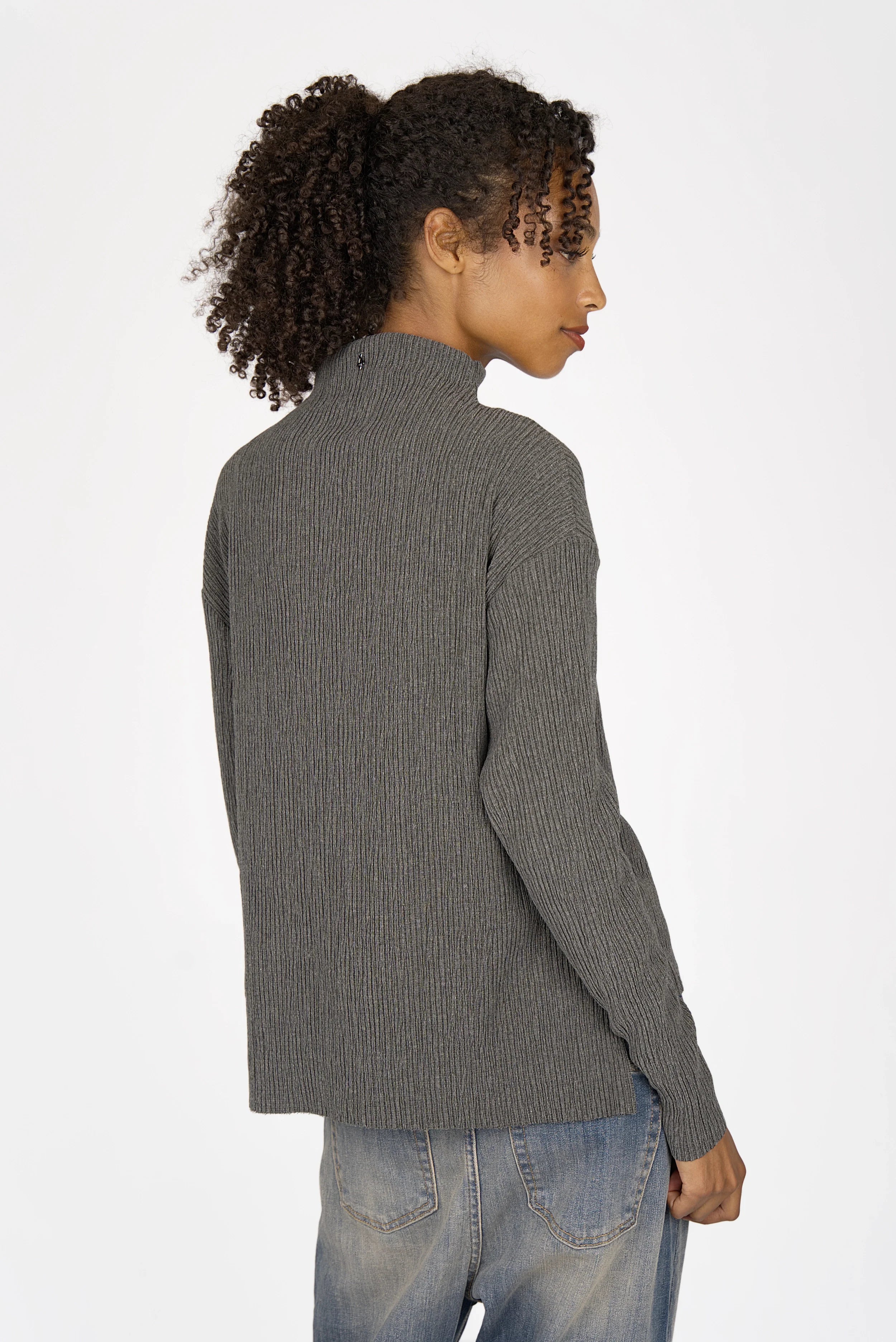 Woman wearing grey NORELI Longsleeve with mock neck and pleated crepe jersey fabric, featuring dropped shoulders and extended back hem with side slits.