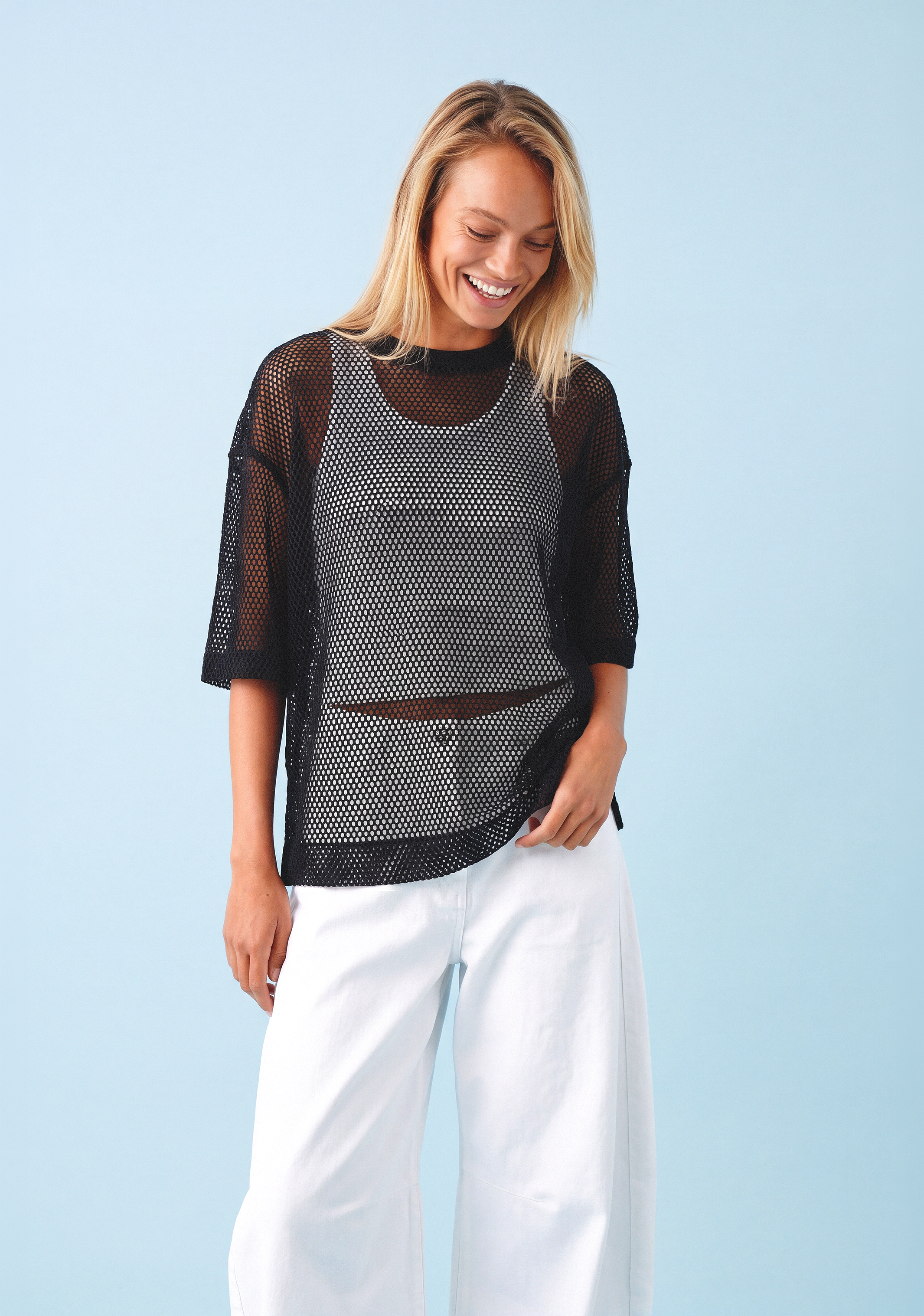 Model im schwarzen Mesh-Top über hellem Top kombiniert mit weiter weißer Hose vor klarem blauem Hintergrund.