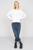 Frau trägt weißes RIISA Longsleeve mit U-Boot-Ausschnitt, überschnittenen Schultern und extra breiten Rippbündchen zu blauen Jeans und schwarzen Stiefeln.