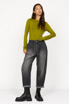 Woman wearing SAIKA Relaxed Tapered Hose in denim-look with gradient wash and whisker folds, paired with olive green long-sleeve top and black boots.