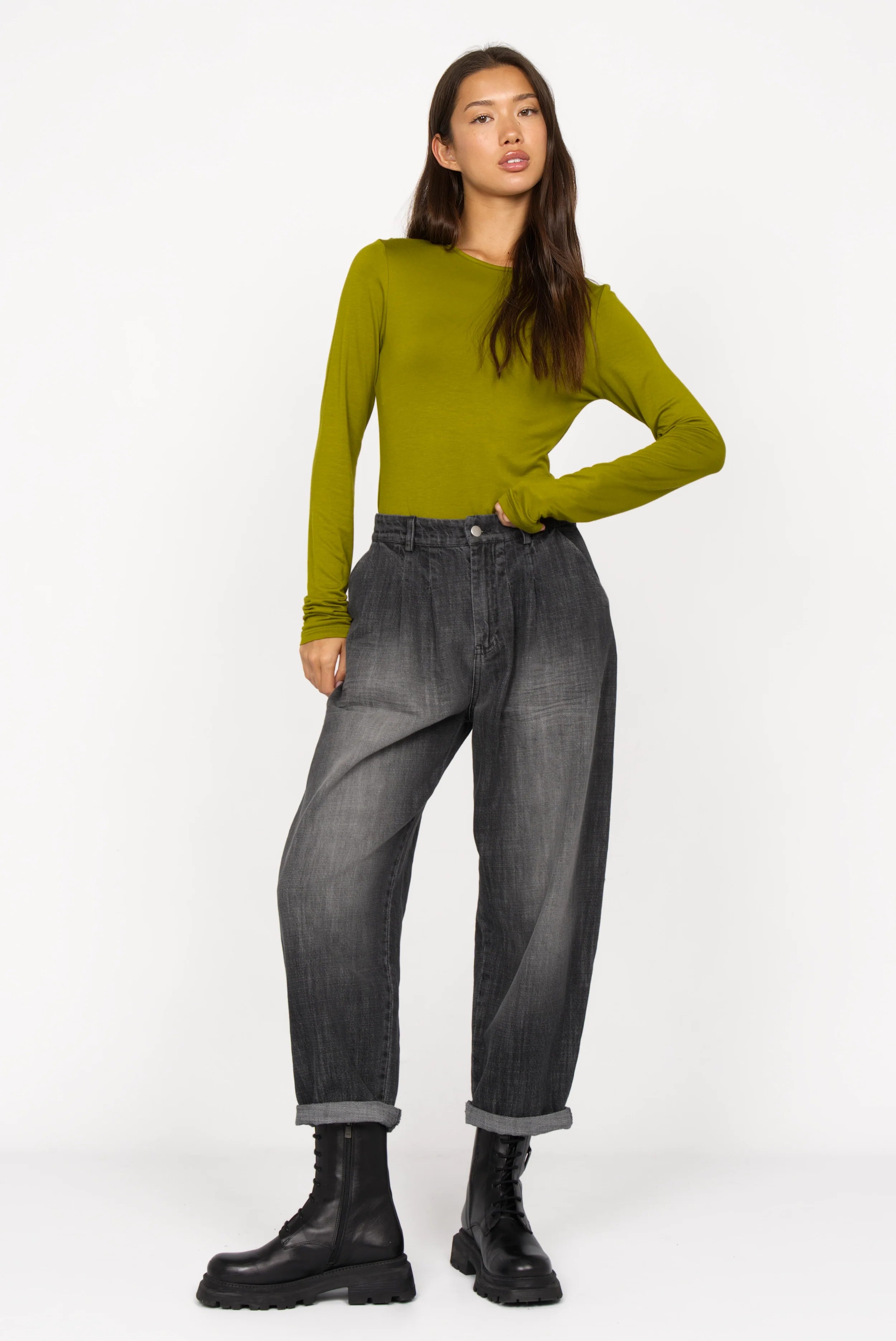 Woman wearing SAIKA Relaxed Tapered Hose in denim-look with gradient wash and whisker folds, paired with olive green long-sleeve top and black boots.