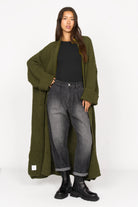 Frau trägt SAIKA Relaxed Tapered Hose in gewaschenem Schwarz mit Whisker-Falten, kombiniert mit langem, grob gestricktem olivgrünem Cardigan und schwarzen Boots.