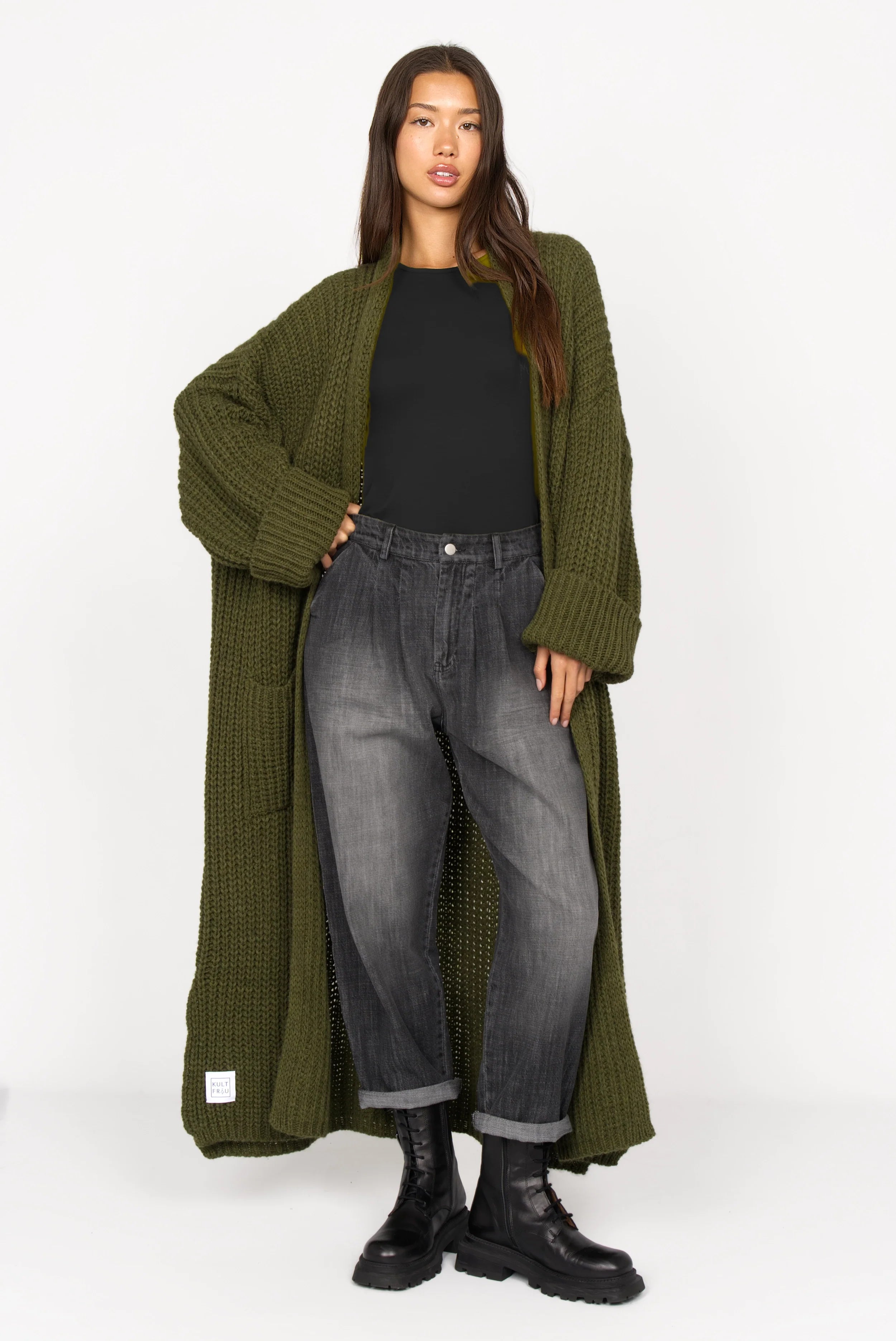 Frau trägt SAIKA Relaxed Tapered Hose in gewaschenem Schwarz mit Whisker-Falten, kombiniert mit langem, grob gestricktem olivgrünem Cardigan und schwarzen Boots.