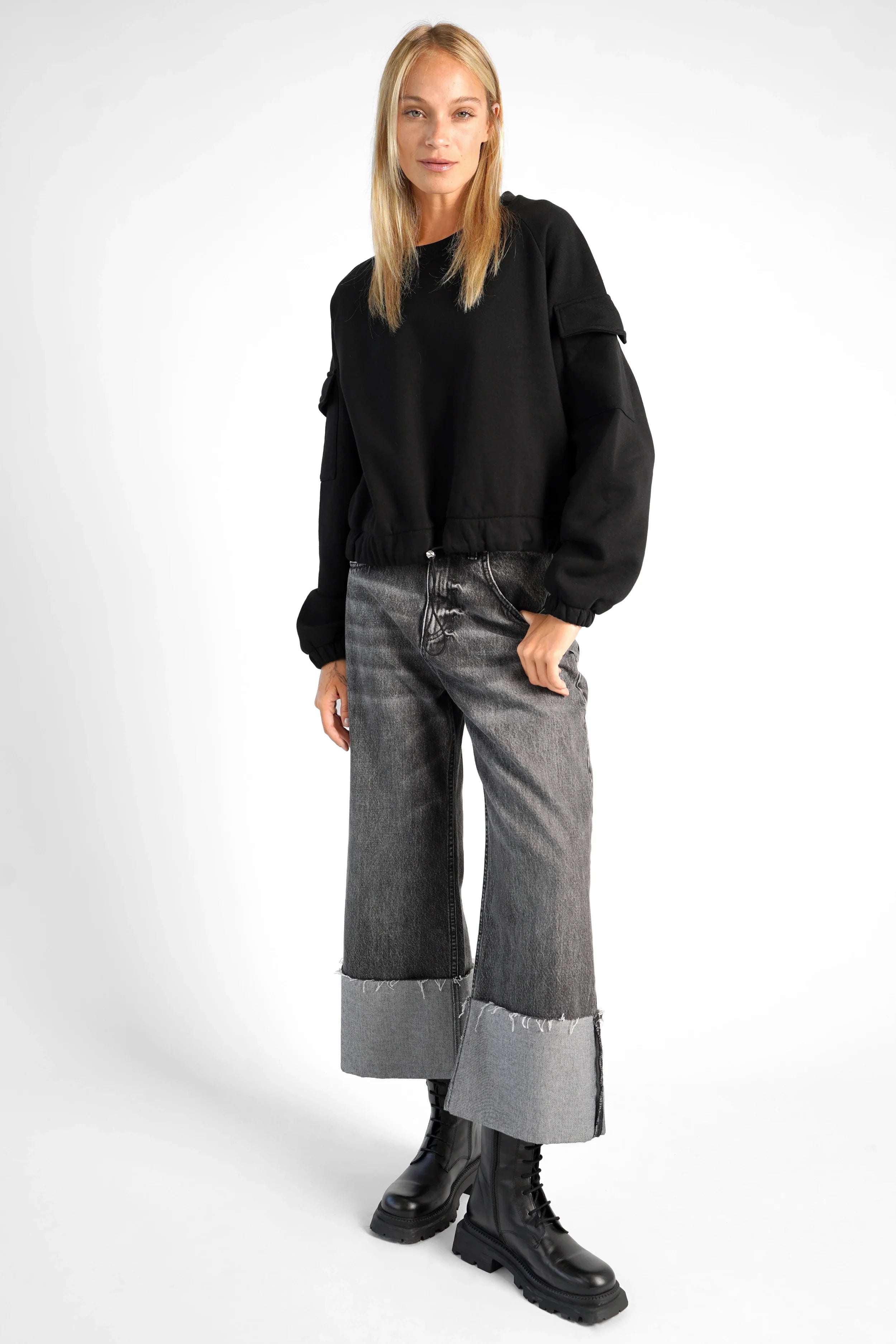 Woman wearing black SEATA Cropped Sweater with oversized fit, round neckline, drawstring waistband, and elastic cuffs paired with wide-leg jeans and black boots