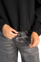 Close-up of a person adjusting the drawstring on a black SEATA Cropped Sweater with elastic cuffs and front pockets, paired with gray denim jeans.