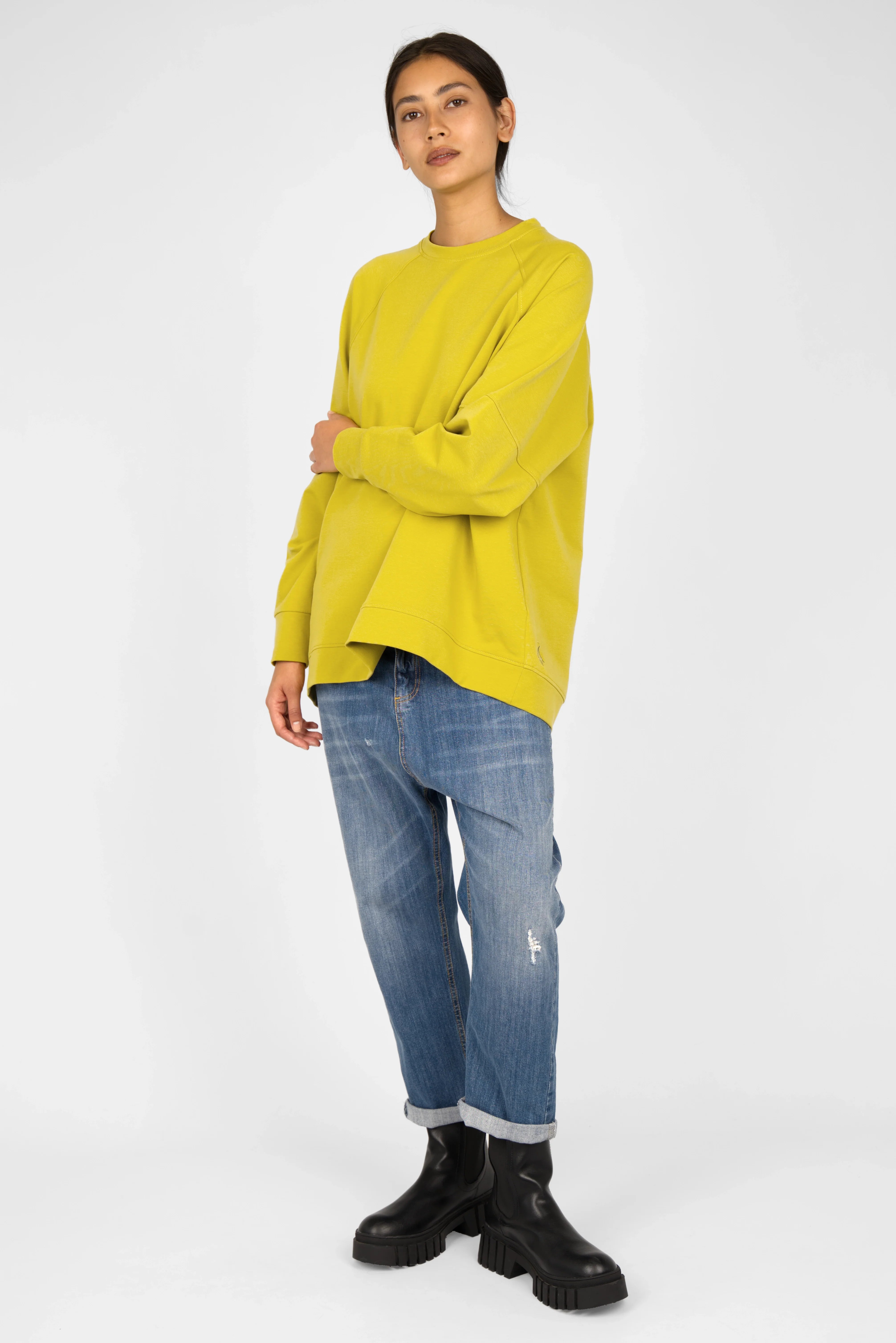 Woman wearing a relaxed fit yellow SELKA Sweater with raglan sleeves and a longer back hem paired with blue rolled-up jeans and black boots.