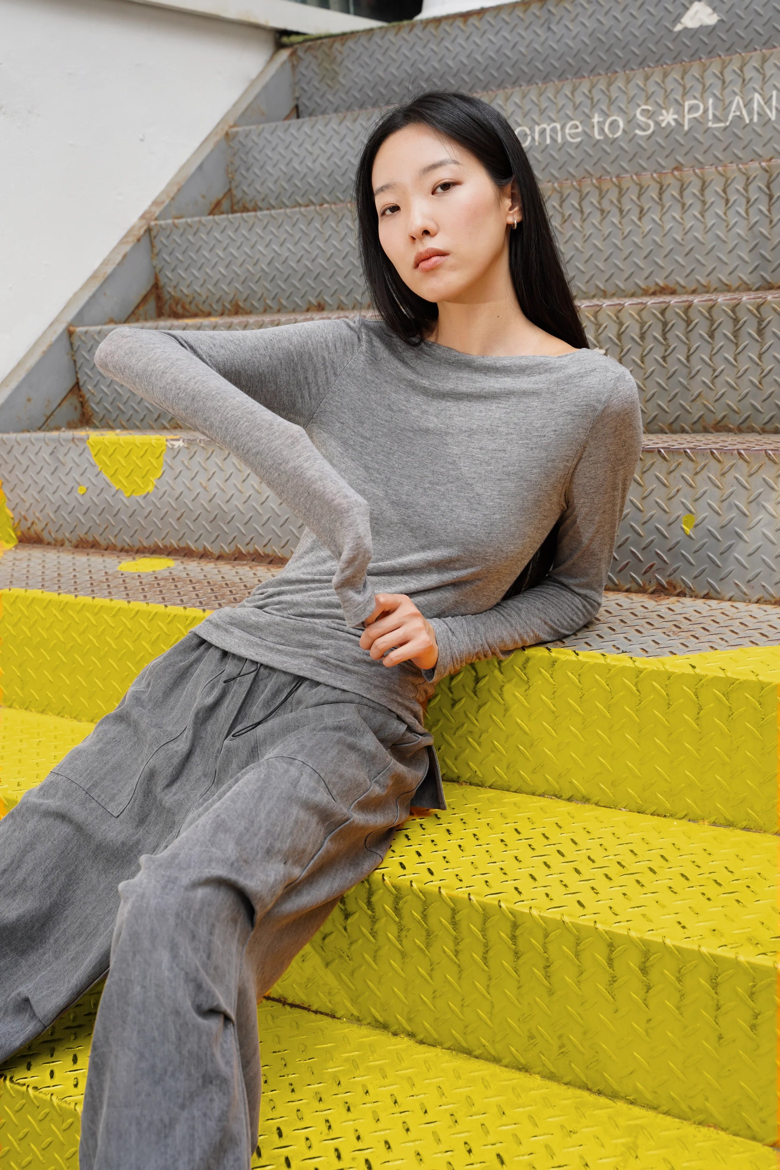Graues SUJIN Longsleeve aus Kaschmirmix mit U-Boot-Ausschnitt und gerafften Ärmeln, slim fit, getragen von Modell auf gelben Metallstufen