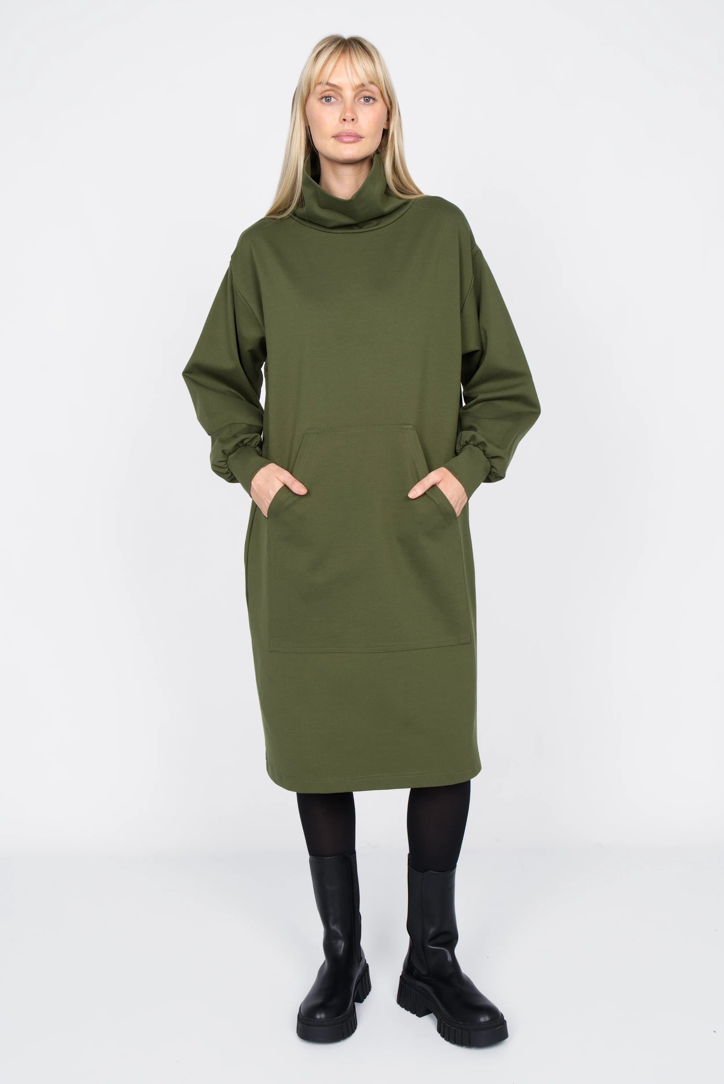 Woman wearing olive green SYNNI Kleid midi dress with loose stand collar and kangaroo pocket, styled with black tights and boots