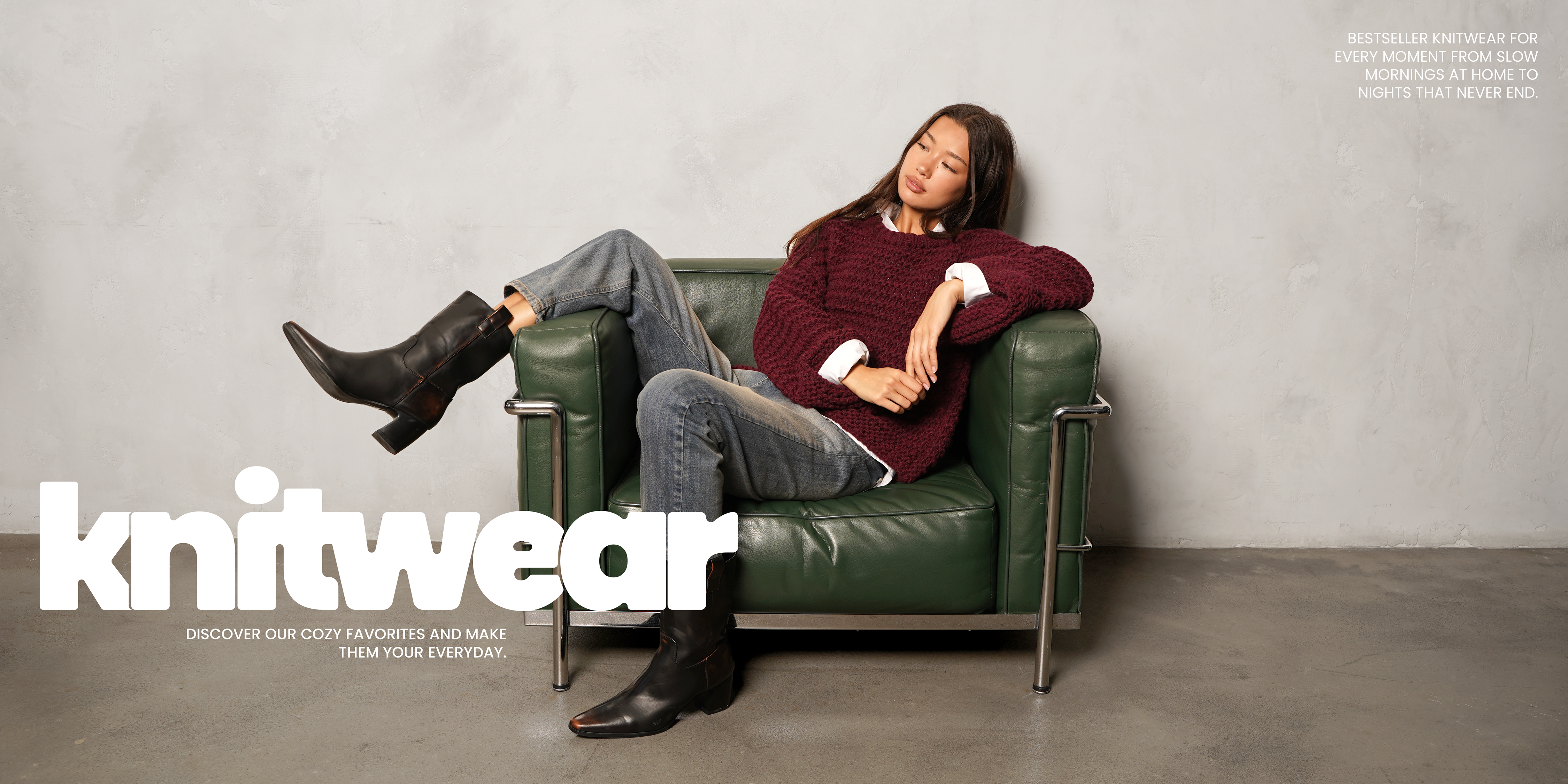 Frau sitzt entspannt auf einem grünen Ledersessel und trägt einen burgunderfarbenen Strickpullover über weißem Hemd, kombiniert mit Jeans und schwarzen Lederstiefeln – gemütlicher, moderner Knitwear-Look.