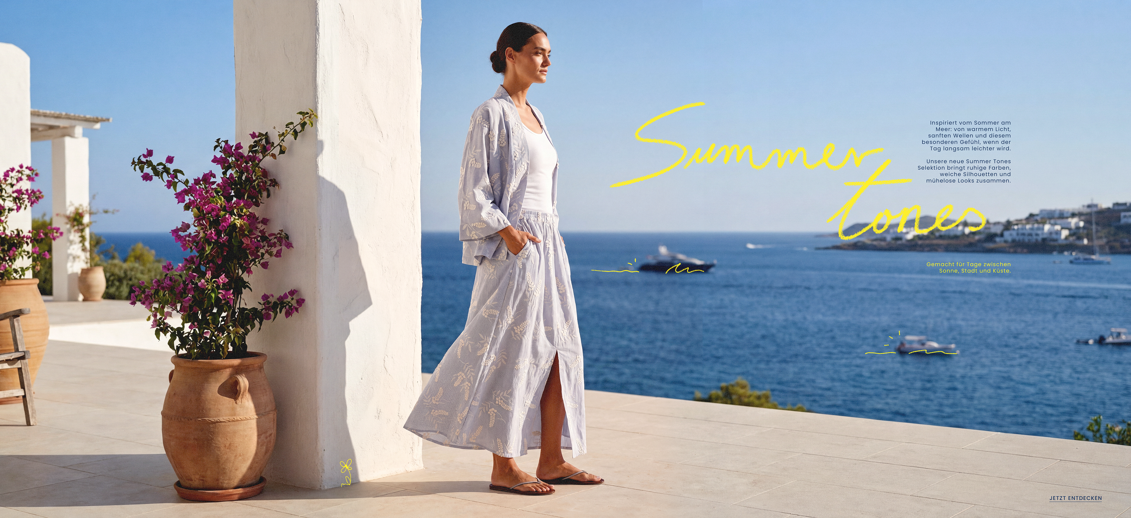 Model in luftigem, hell gemustertem Zweiteiler mit langem Rock und Kimono-Jacke auf mediterraner Terrasse mit Meerblick – Sommerkollektion in sanften Tönen.