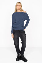 Woman wearing blue asymmetrical one-shoulder pleated crepe jersey top with wide batwing sleeve and raw-cut hem, paired with black pants and boots