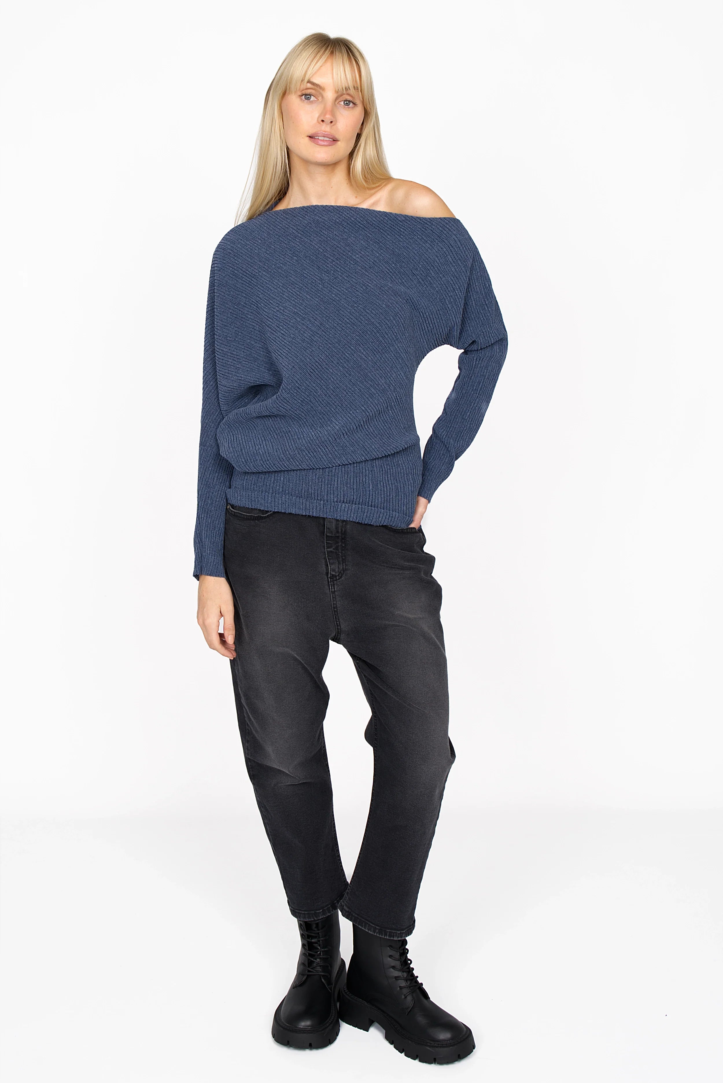 Woman wearing blue asymmetrical one-shoulder pleated crepe jersey top with wide batwing sleeve and raw-cut hem, paired with black pants and boots
