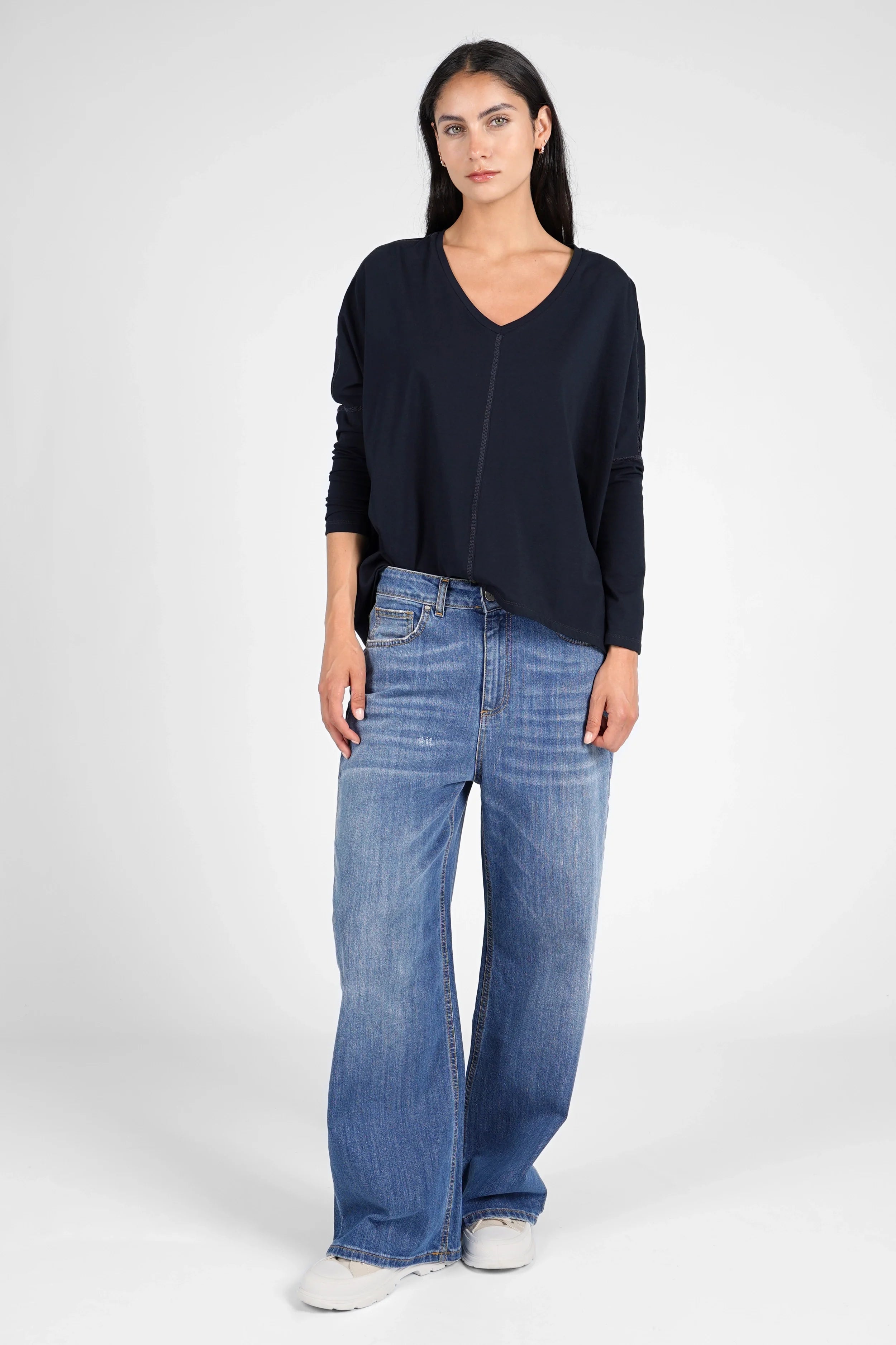 Woman wearing a navy TIDA Blusenshirt long sleeve shirt with V-neck and relaxed boxy fit, paired with wide-leg blue jeans and white sneakers.