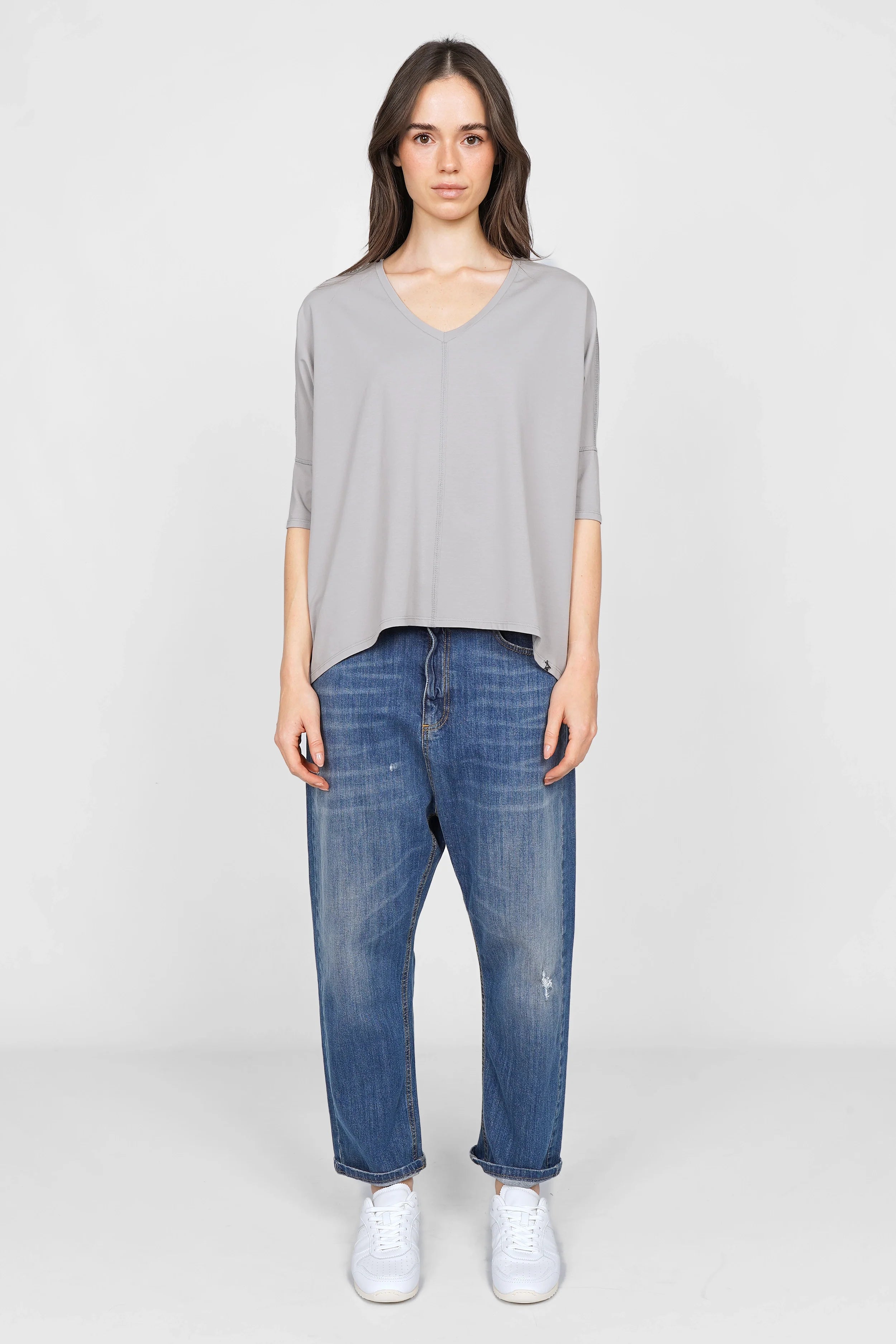Damen trägt graues TIDA Blusenshirt mit V-Ausschnitt und weiter Kastenform zu blauen Jeans und weißen Sneakern