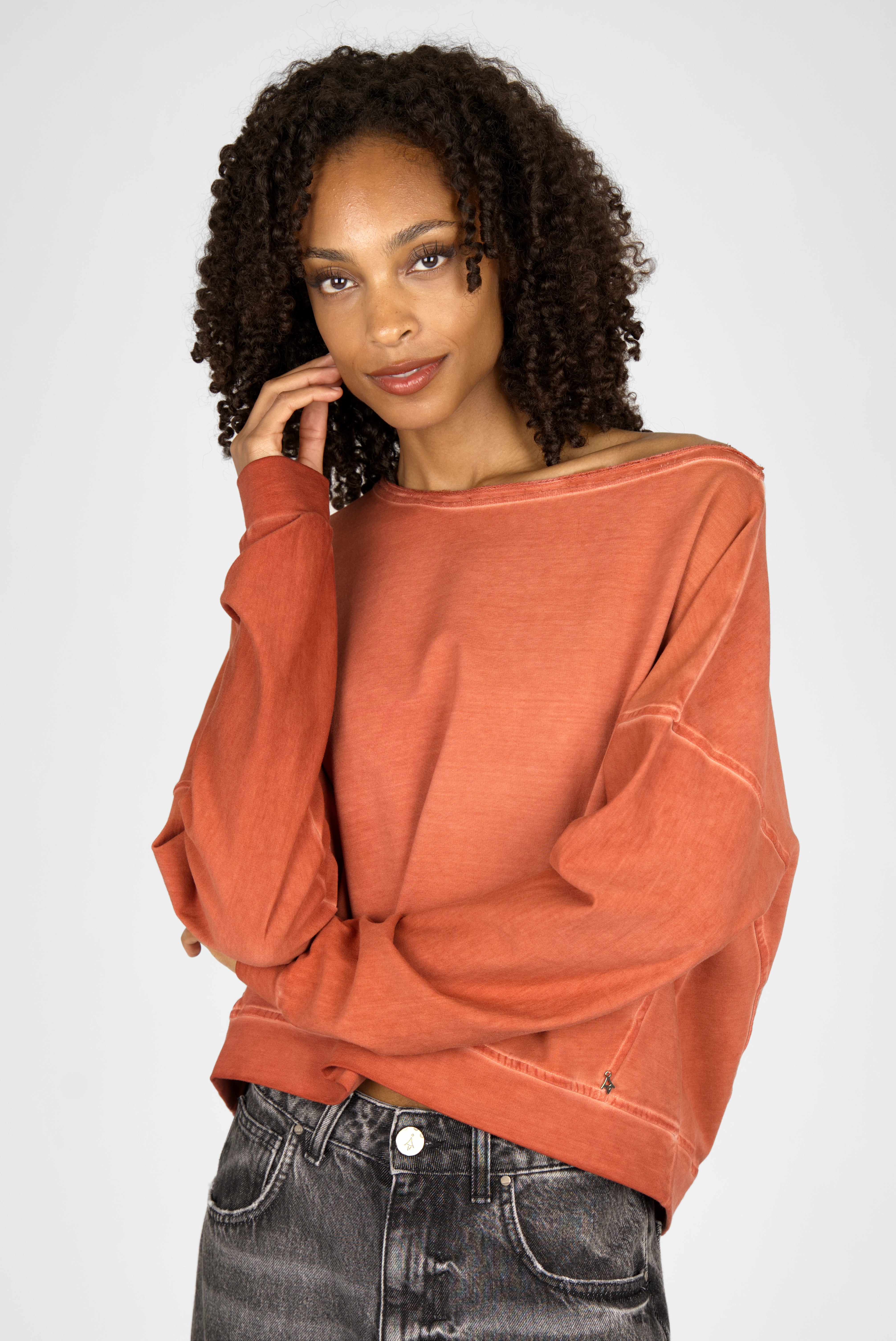 Woman wearing TIMAJ Revive One Shoulder Sweater in relaxed fit with wide neckline and raw edges in rust color.