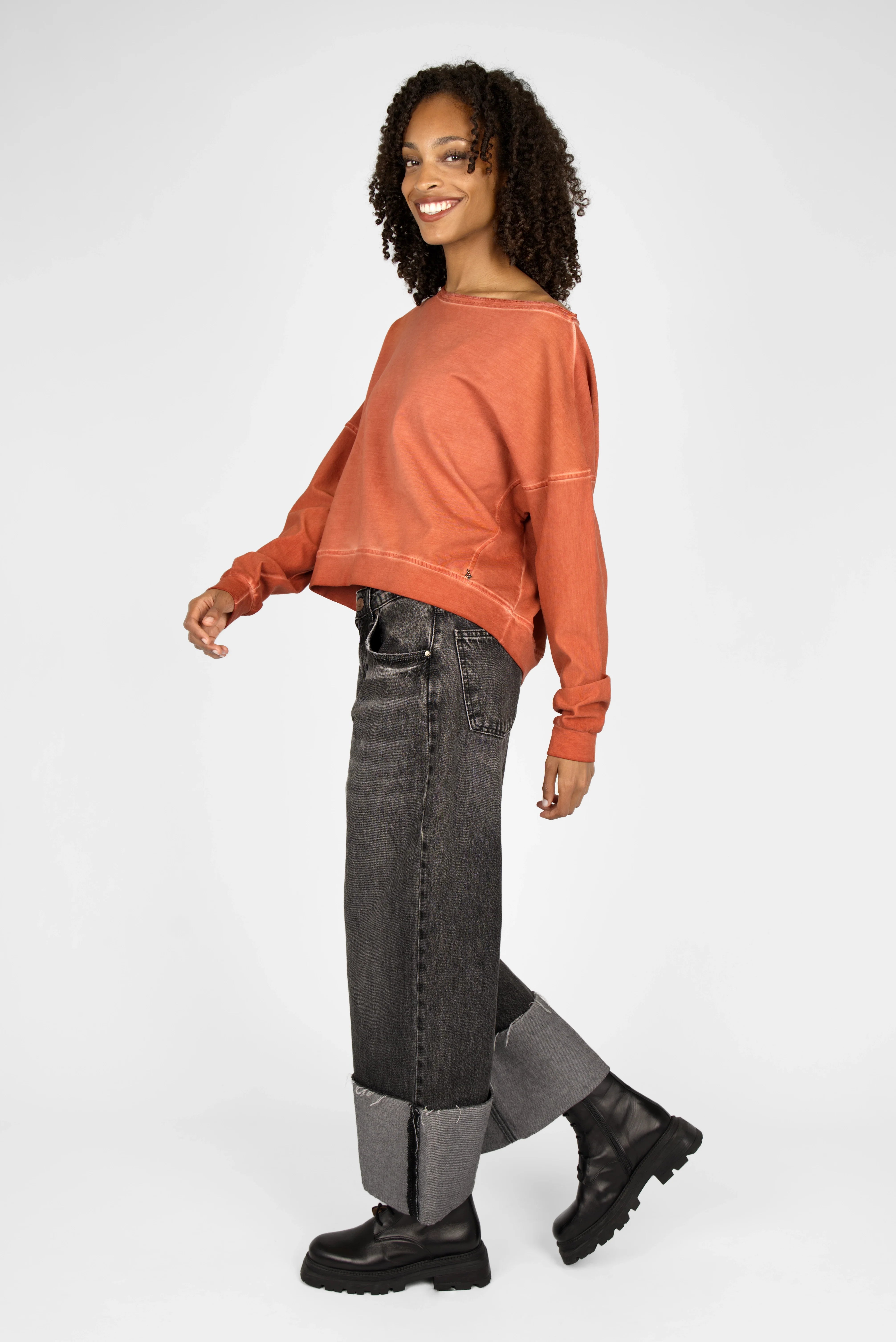 Woman wearing a rust-colored TIMAJ Revive One Shoulder Sweater with loose fit and wide neckline paired with dark rolled-up jeans and black boots.