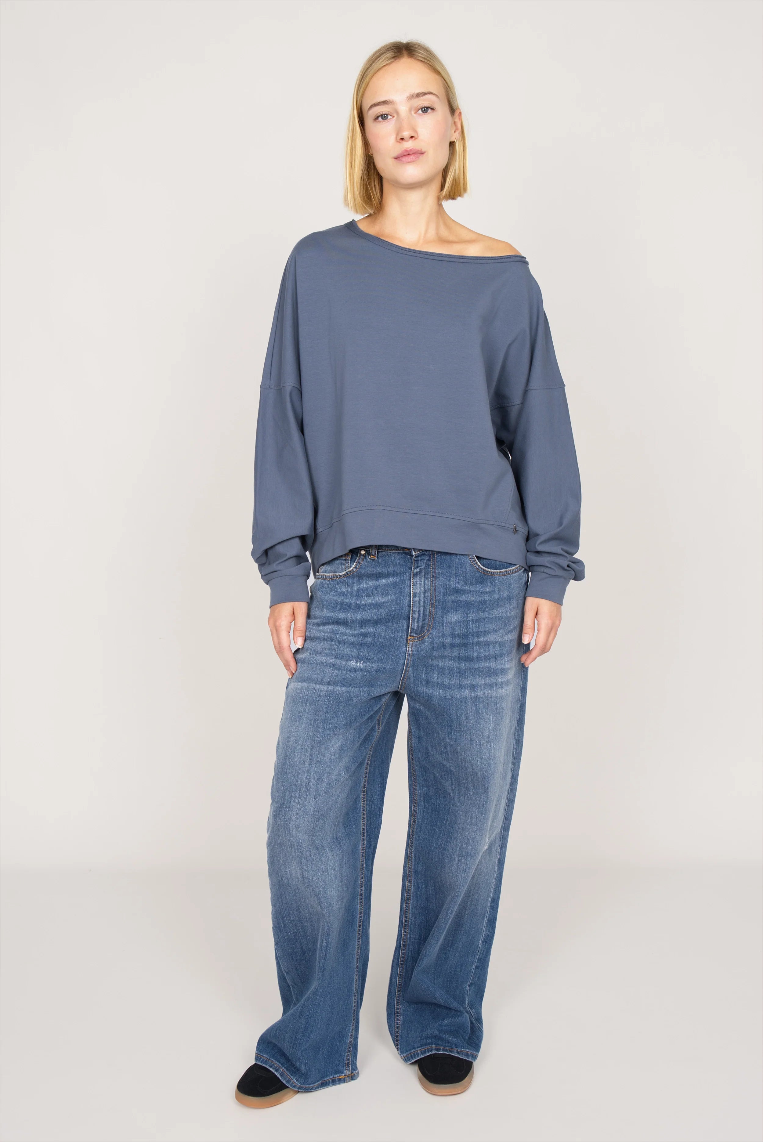 Blauer One Shoulder Sweater aus weichem Sweatstoff mit weitem Halsausschnitt und lässigem Loose Fit, kombiniert mit weiten Jeans.