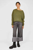 Woman wearing an olive green TIMAJ Revive One Shoulder Sweater with loose fit, wide neckline, and open edges paired with wide-leg cuffed jeans and black boots.