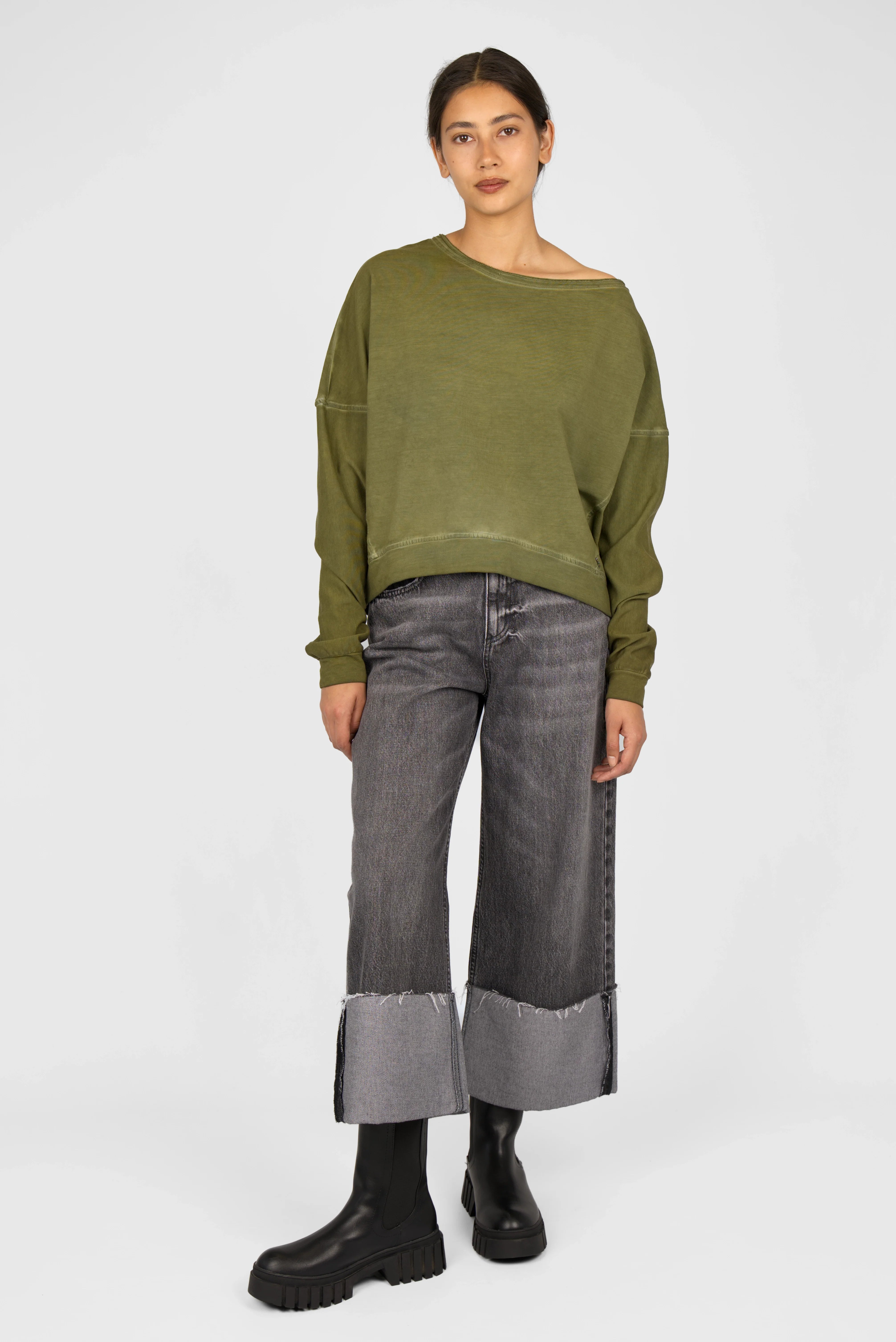 Woman wearing an olive green TIMAJ Revive One Shoulder Sweater with loose fit, wide neckline, and open edges paired with wide-leg cuffed jeans and black boots.