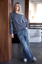 Frau trägt grauen One Shoulder Sweater mit weitem Halsausschnitt und offenen Kanten kombiniert mit weiten Jeans in modernem Wohnzimmer