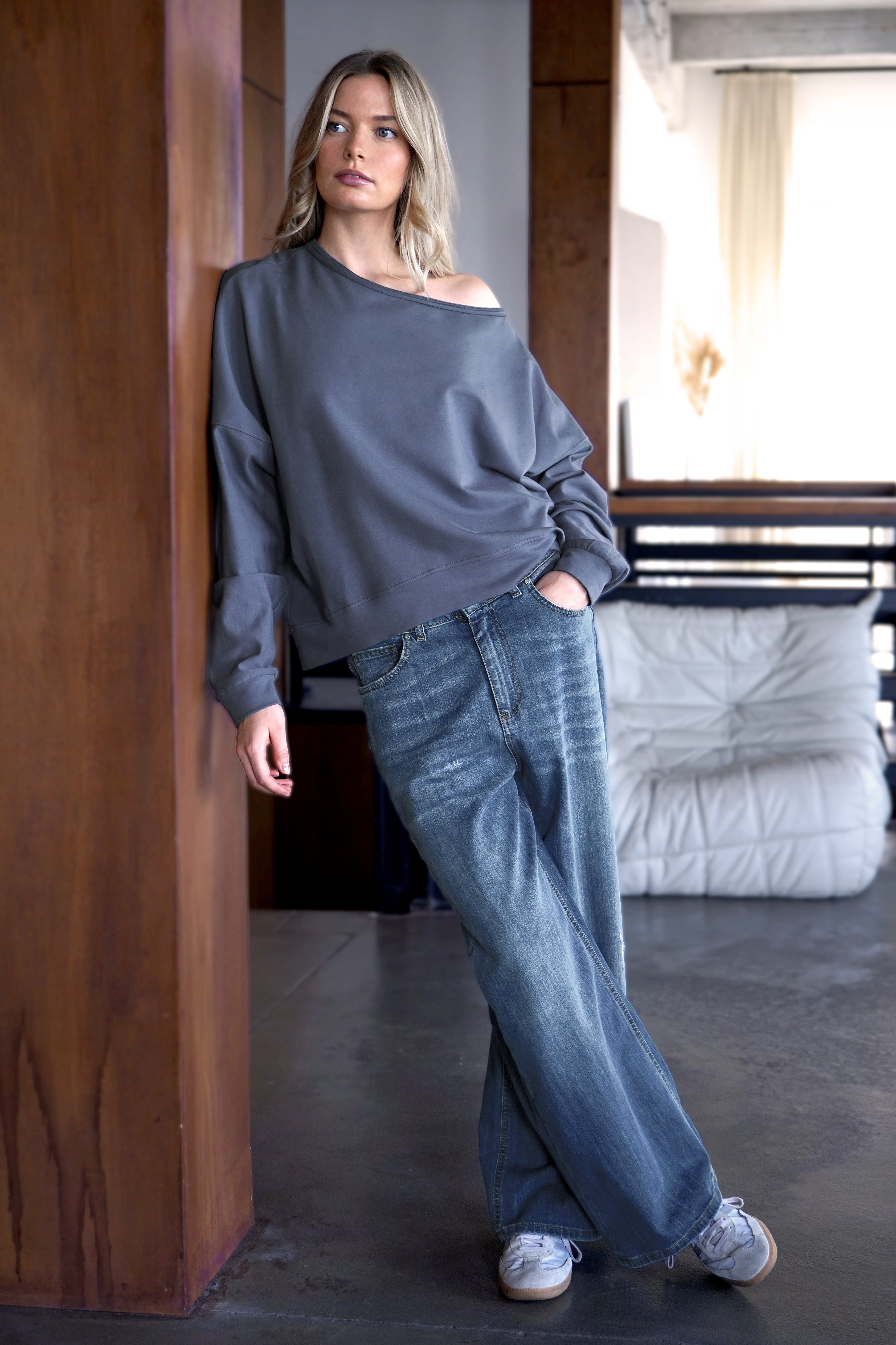 Frau trägt grauen One Shoulder Sweater mit weitem Halsausschnitt und offenen Kanten kombiniert mit weiten Jeans in modernem Wohnzimmer