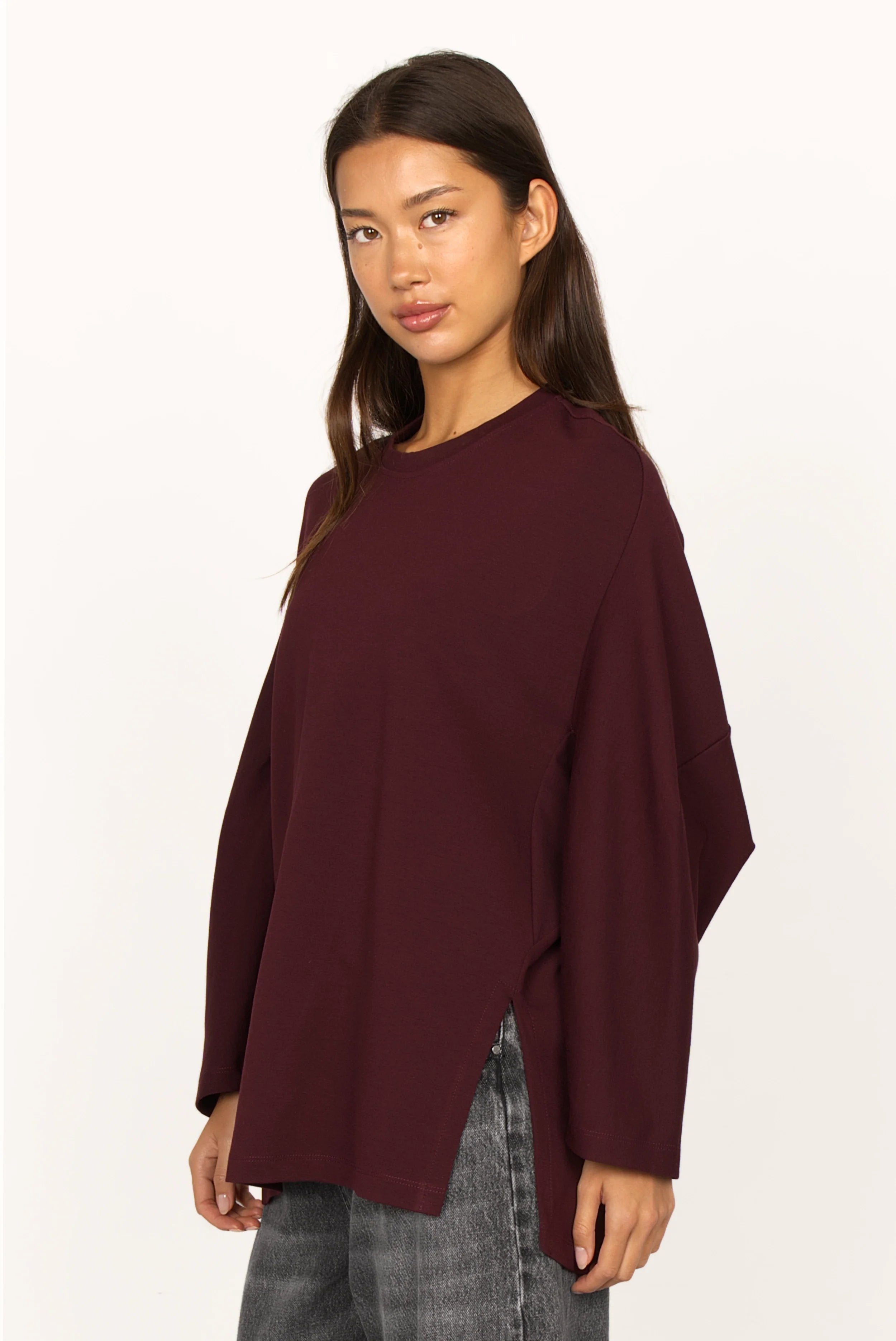 Woman wearing a relaxed fit TOOVE Shirt in dark burgundy with long sleeves, curved seam details, high side slits, and extended back length.